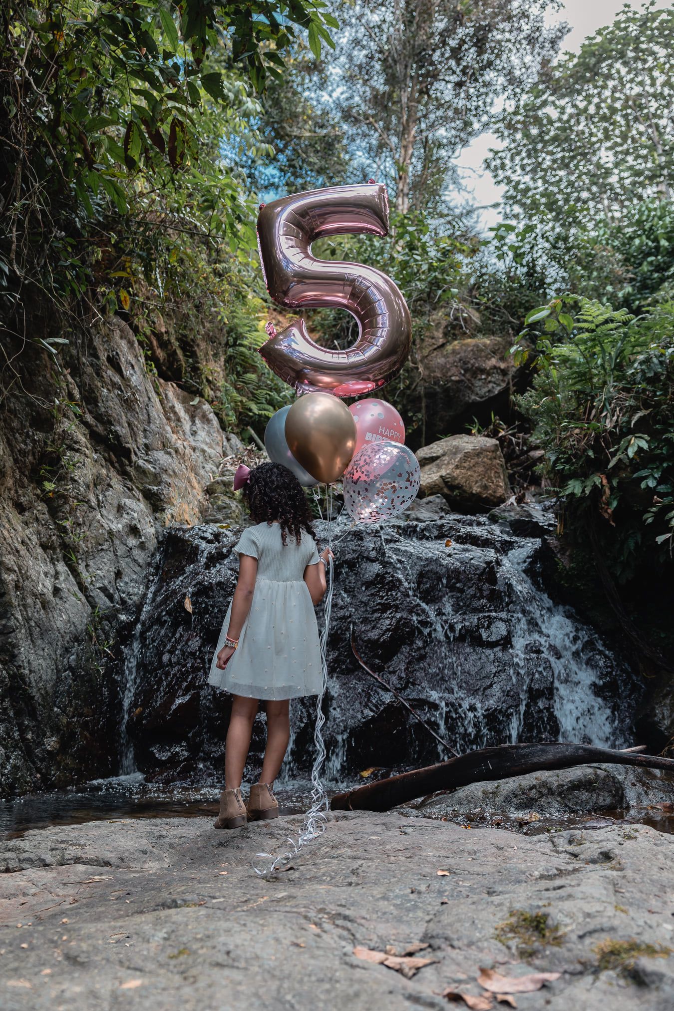 Birthday portrait of a girl holding number five and colorful balloons beside a tropical waterfall