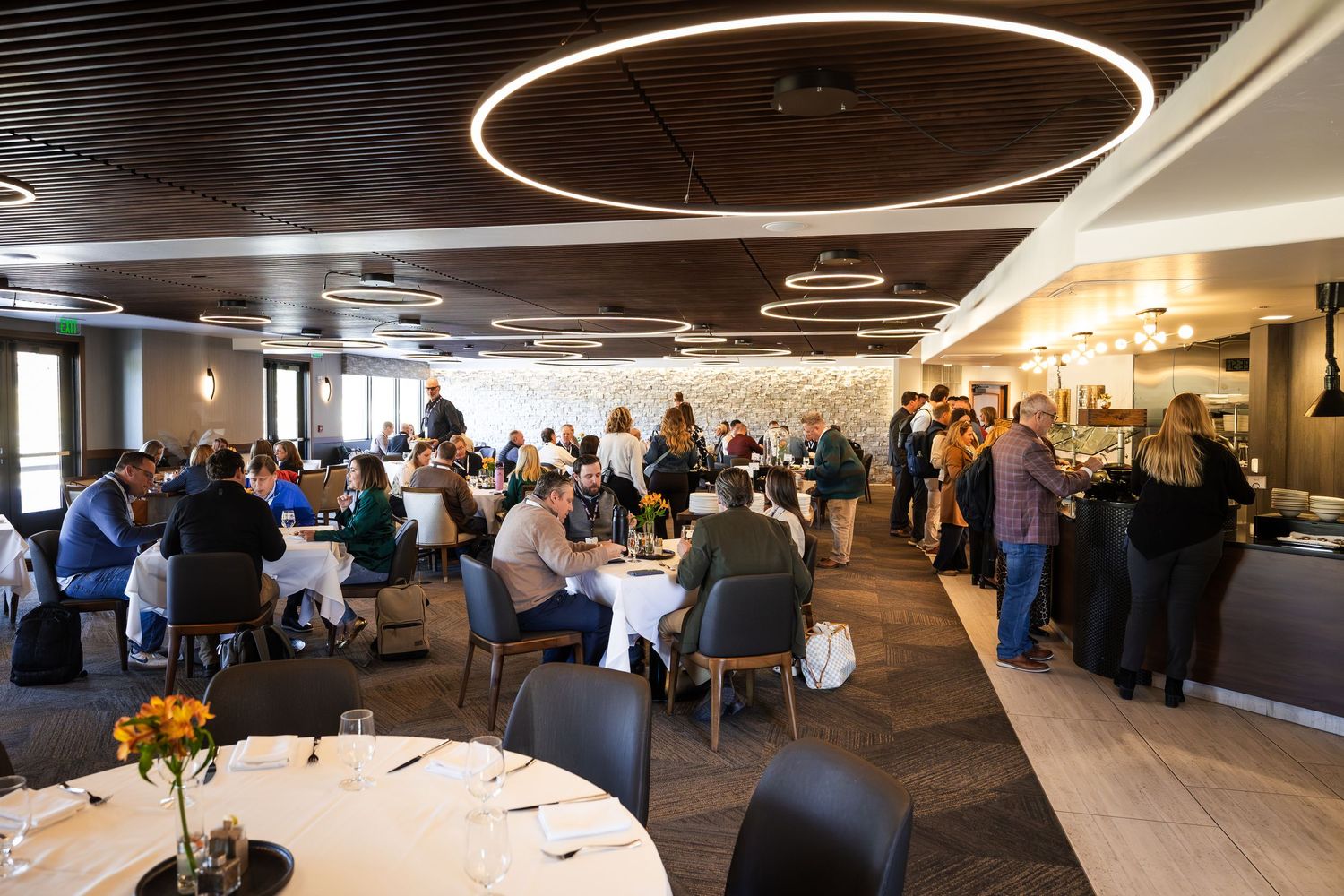 Busy upscale restaurant dining room with circular pendant lights, white tablecloths, and guests enjoying meals.