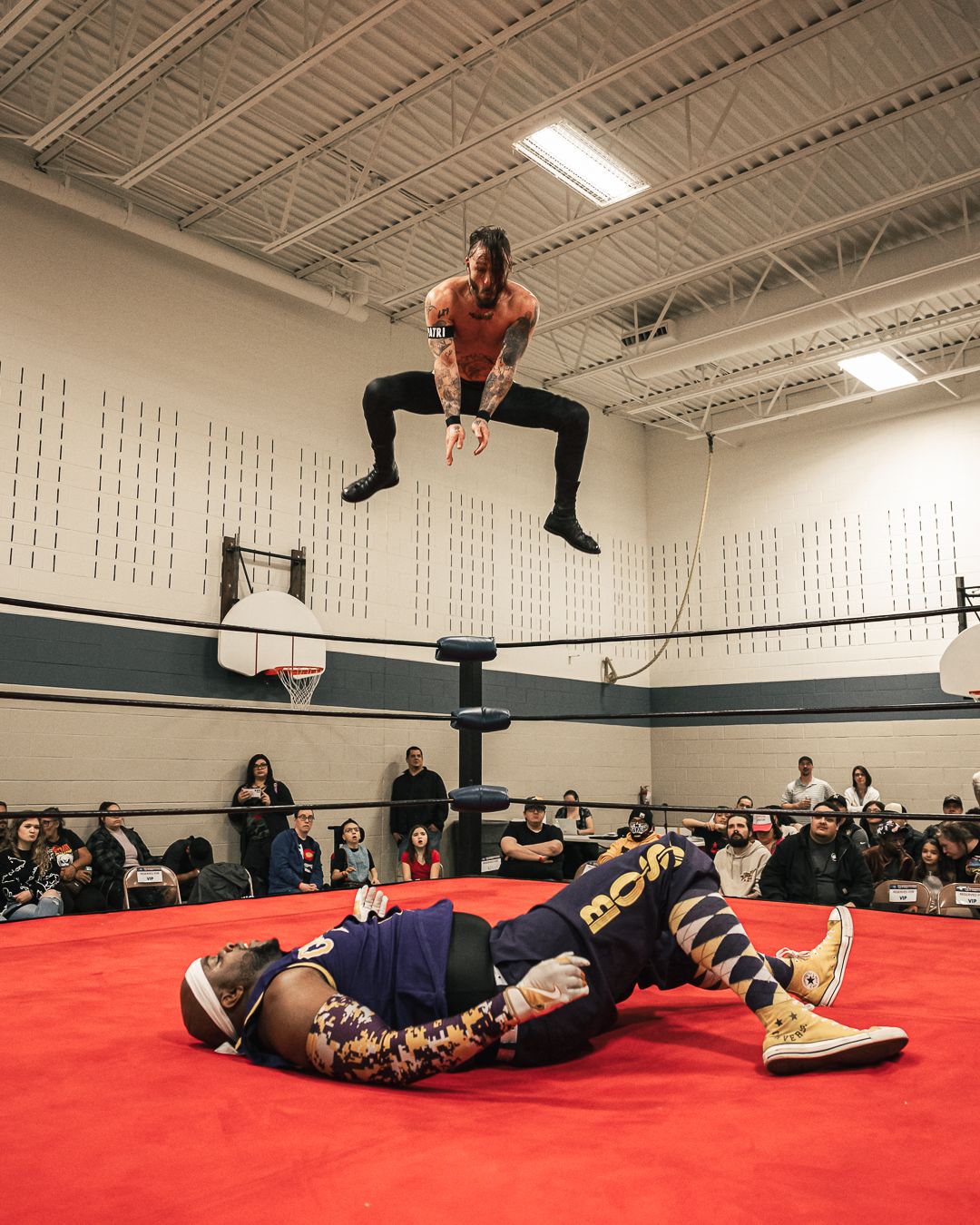 A wrestler leaps high above an opponent lying on a red mat in a gymnasium ring, with spectators watching in the background.