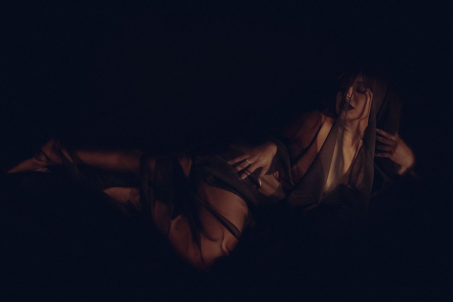 A woman with long black transparent cover reclines in dramatic low-key lighting against a dark background.