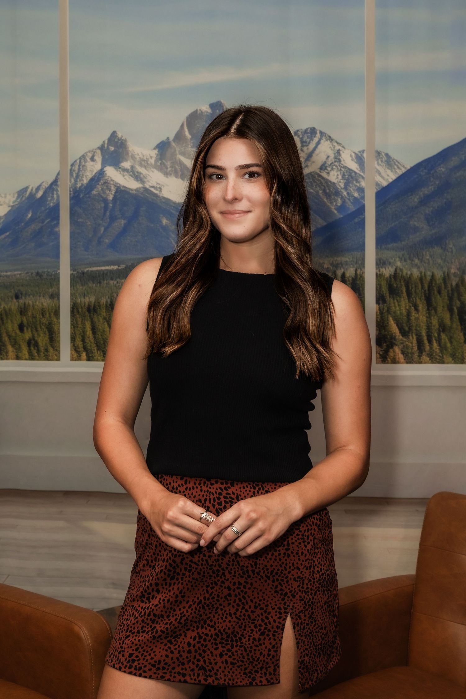 Young woman in black top and leopard print skirt poses indoors with mountain landscape backdrop.
