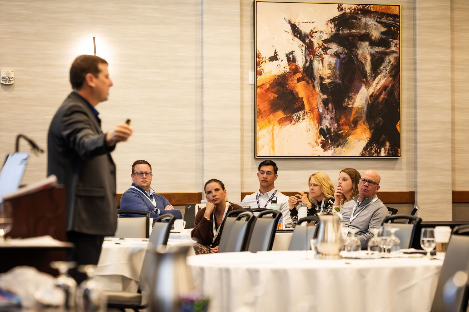 Business professional presenting to engaged audience at corporate conference with abstract horse painting in background.