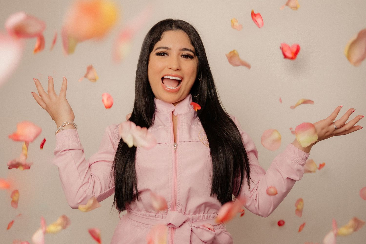 Young woman in pink outfit joyfully posing with rose petals falling around her against a neutral background.