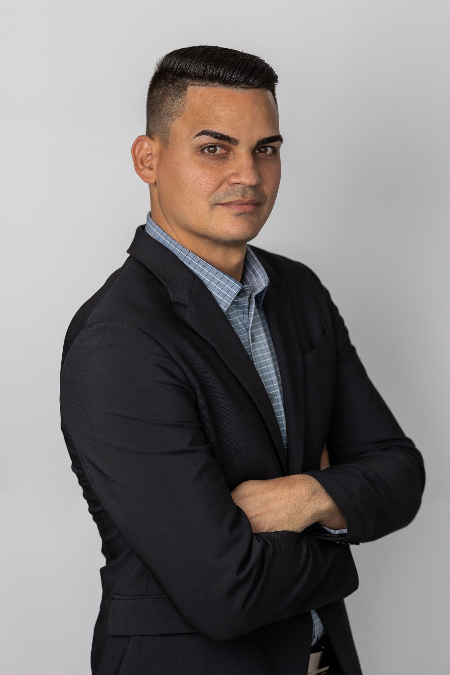 Professional man in black blazer and blue shirt stands with arms crossed against a neutral gray background.