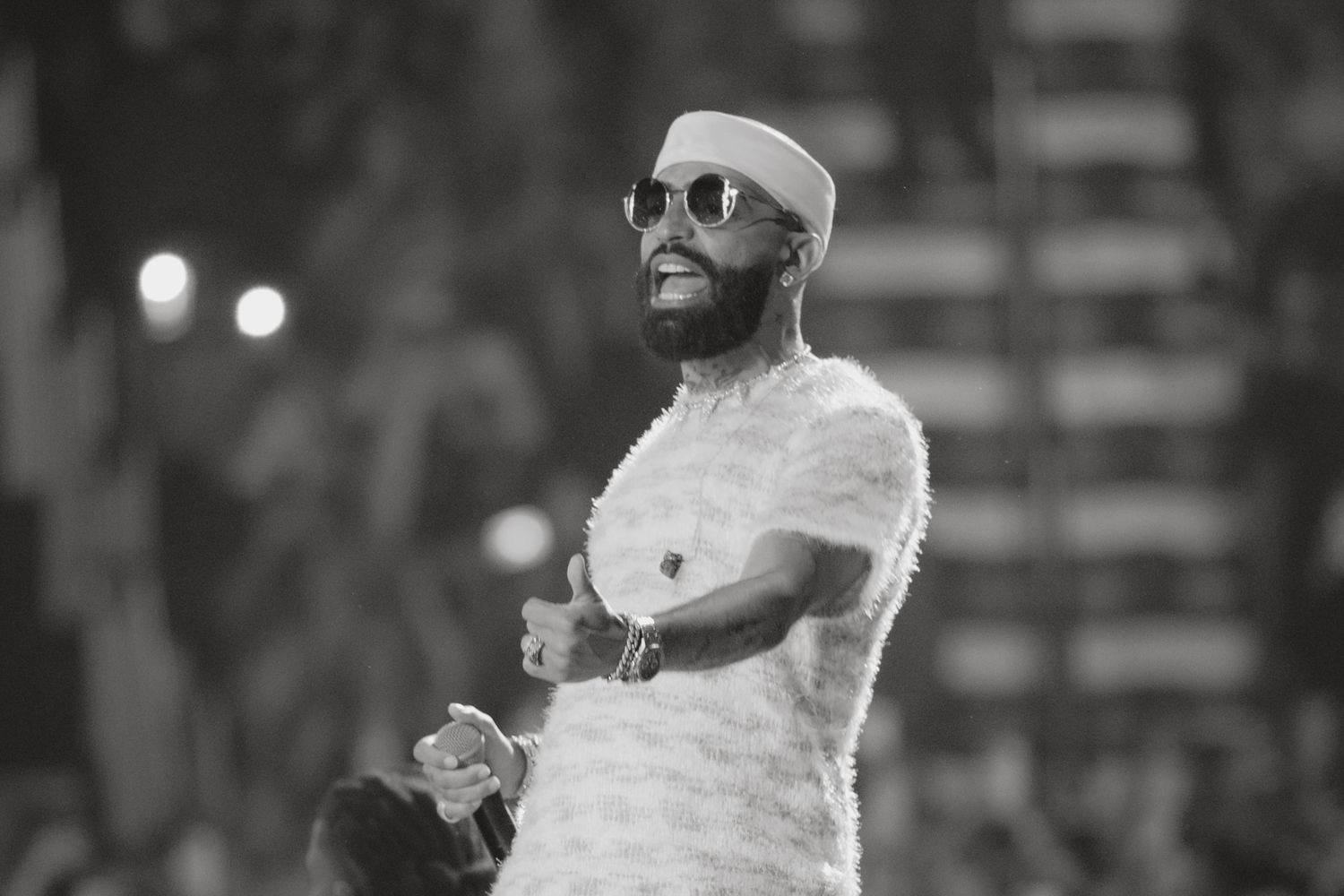 Man in white feathered outfit and sunglasses performing on stage in black and white photo.