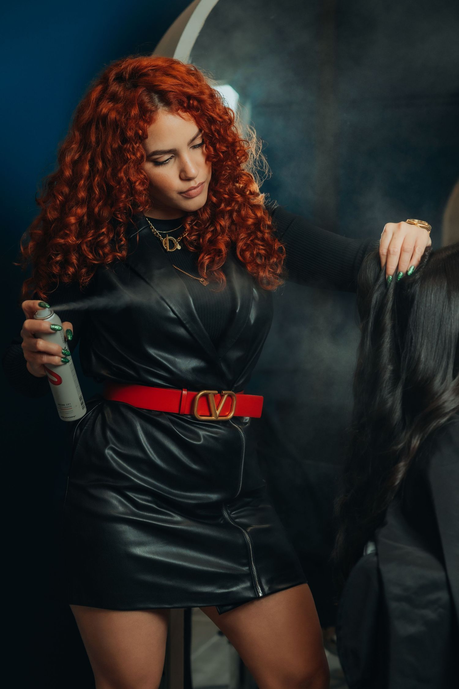 Red curly haired woman in black leather dress with red Valentino belt spraying hairspray on long dark hair.