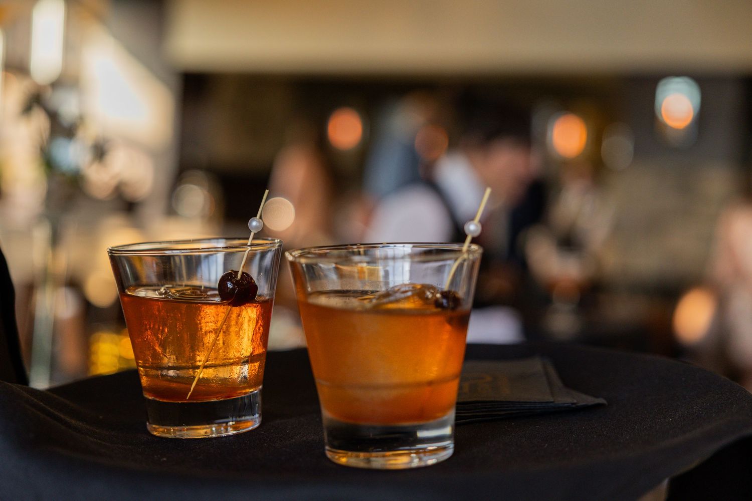 Two whiskey cocktails on the rocks in crystal glasses garnished with cherries, served on a dark bar surface.