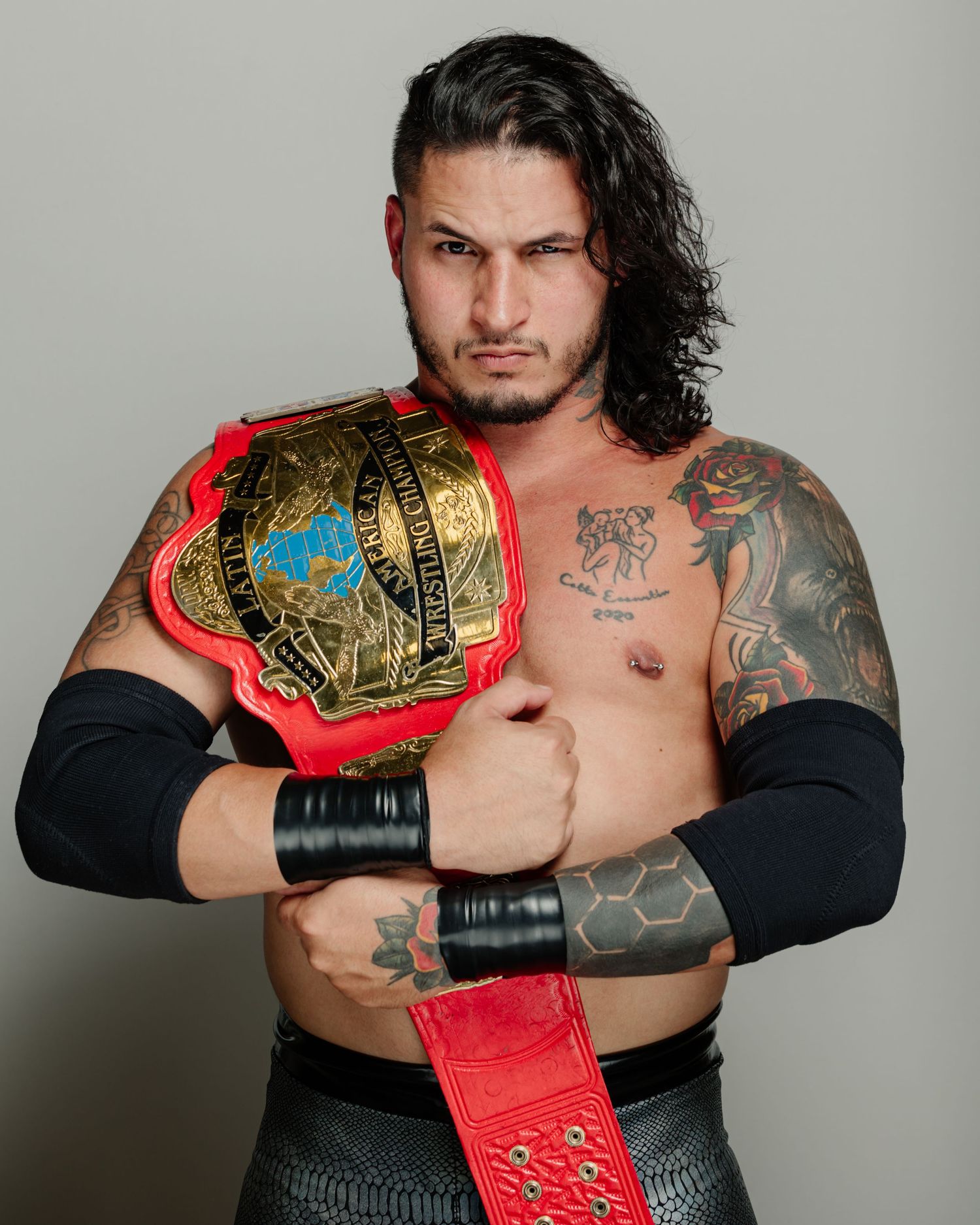 Muscular male wrestler with long dark hair and tattoos posing with a red and gold championship wrestling belt.