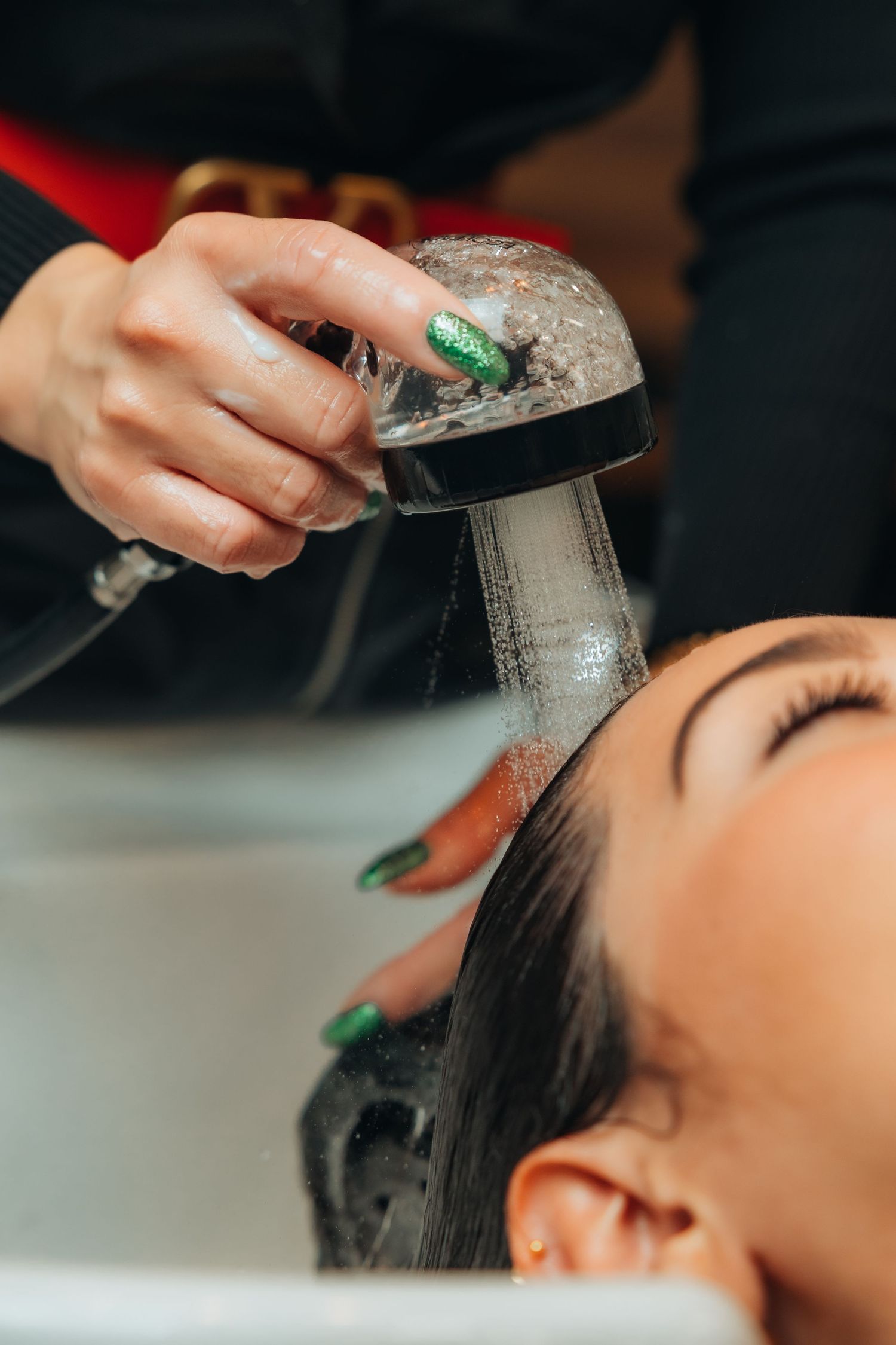 Hairstylist with green nails rinsing dark hair at salon sink using a shower head sprayer.