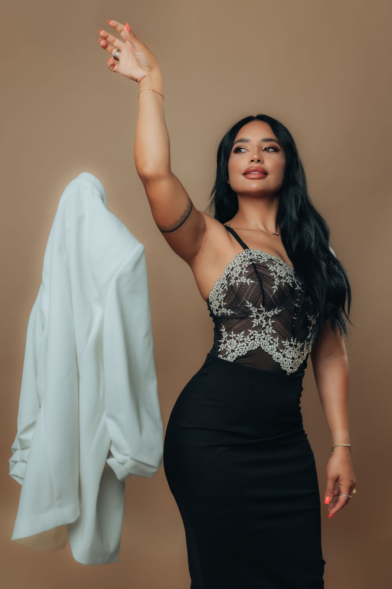 Woman in black lace bodysuit and skirt raises white jacket overhead, posing against a tan background.