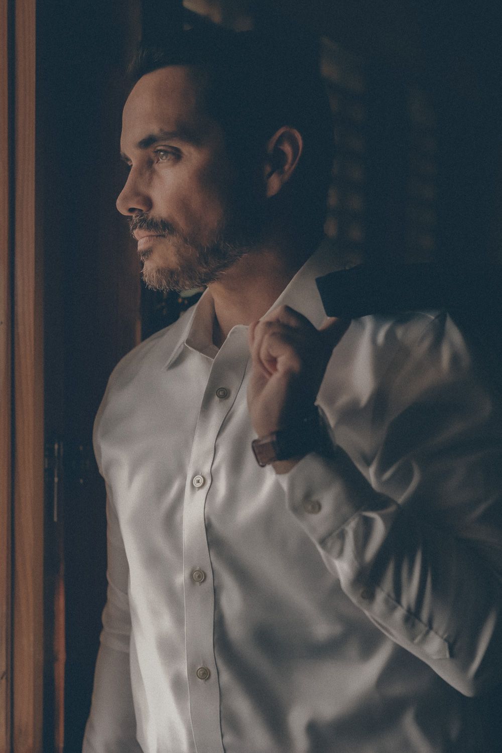 A well-dressed man in a white shirt adjusts his jacket collar while gazing thoughtfully out a window in dim light.