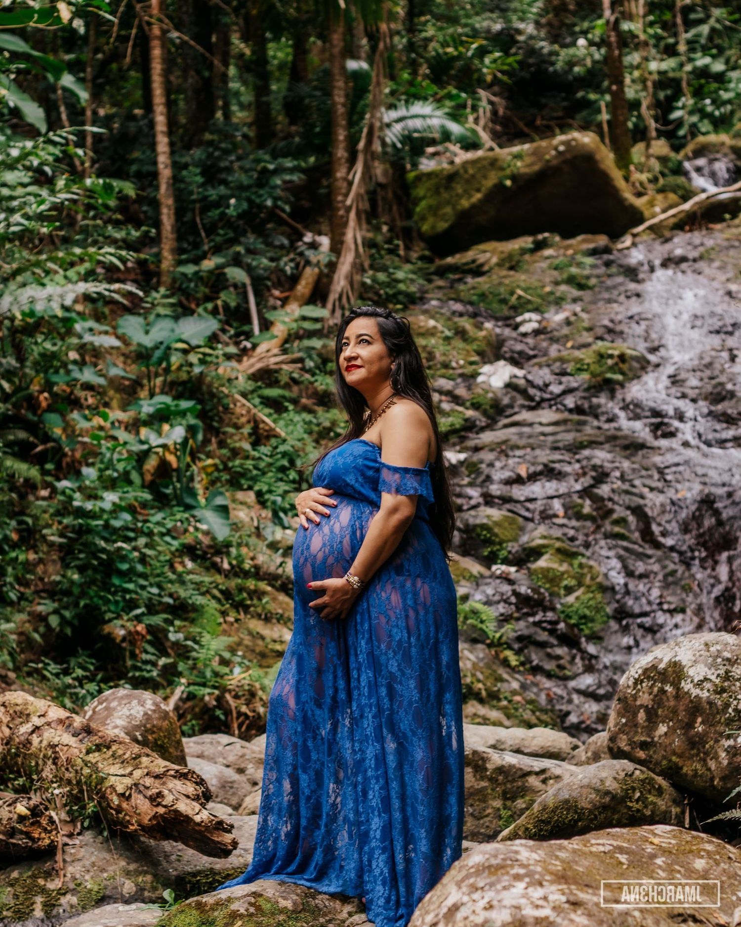 Expecting mother in a blue lace maternity gown posing beside a tropical waterfall