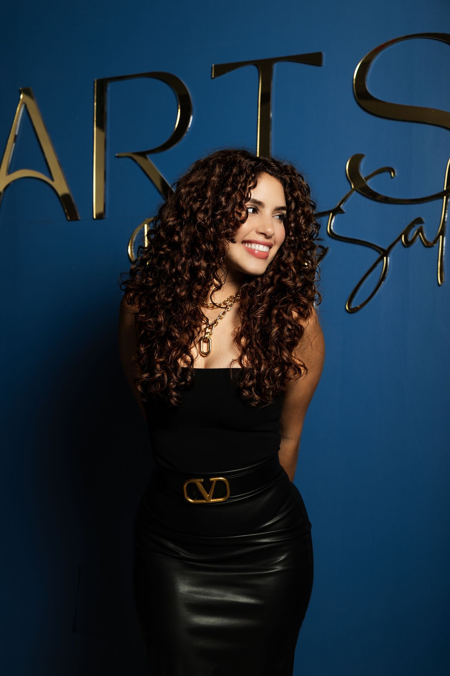 Woman with long curly hair wearing black strapless top and leather skirt with Valentino belt poses by blue wall.
