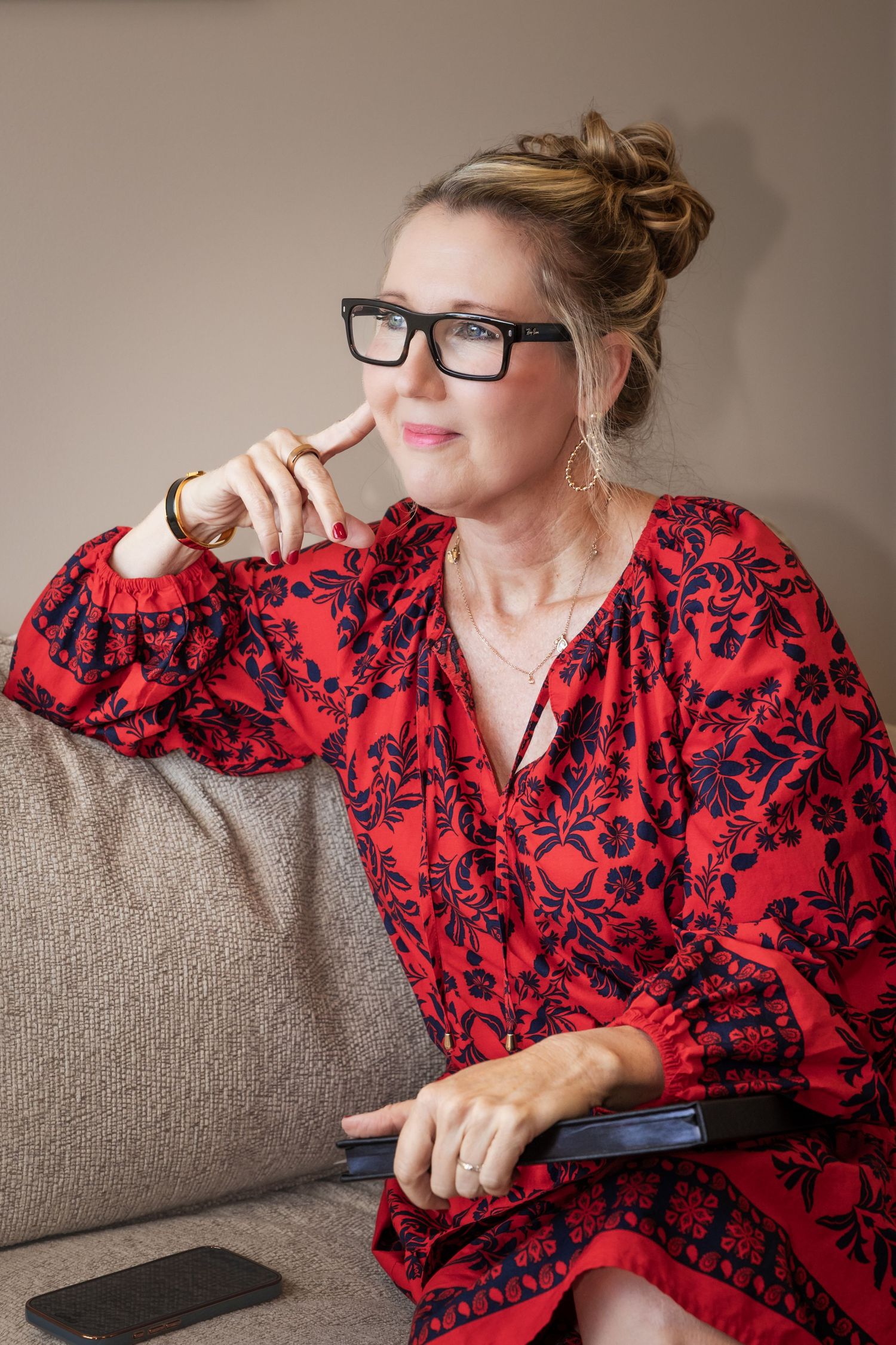 Woman in red floral dress and black glasses sitting thoughtfully on a sofa, holding a tablet.