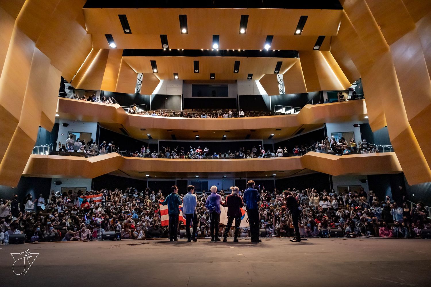 A packed multi-tiered concert hall audience faces performers on stage in a modern wood-paneled venue.