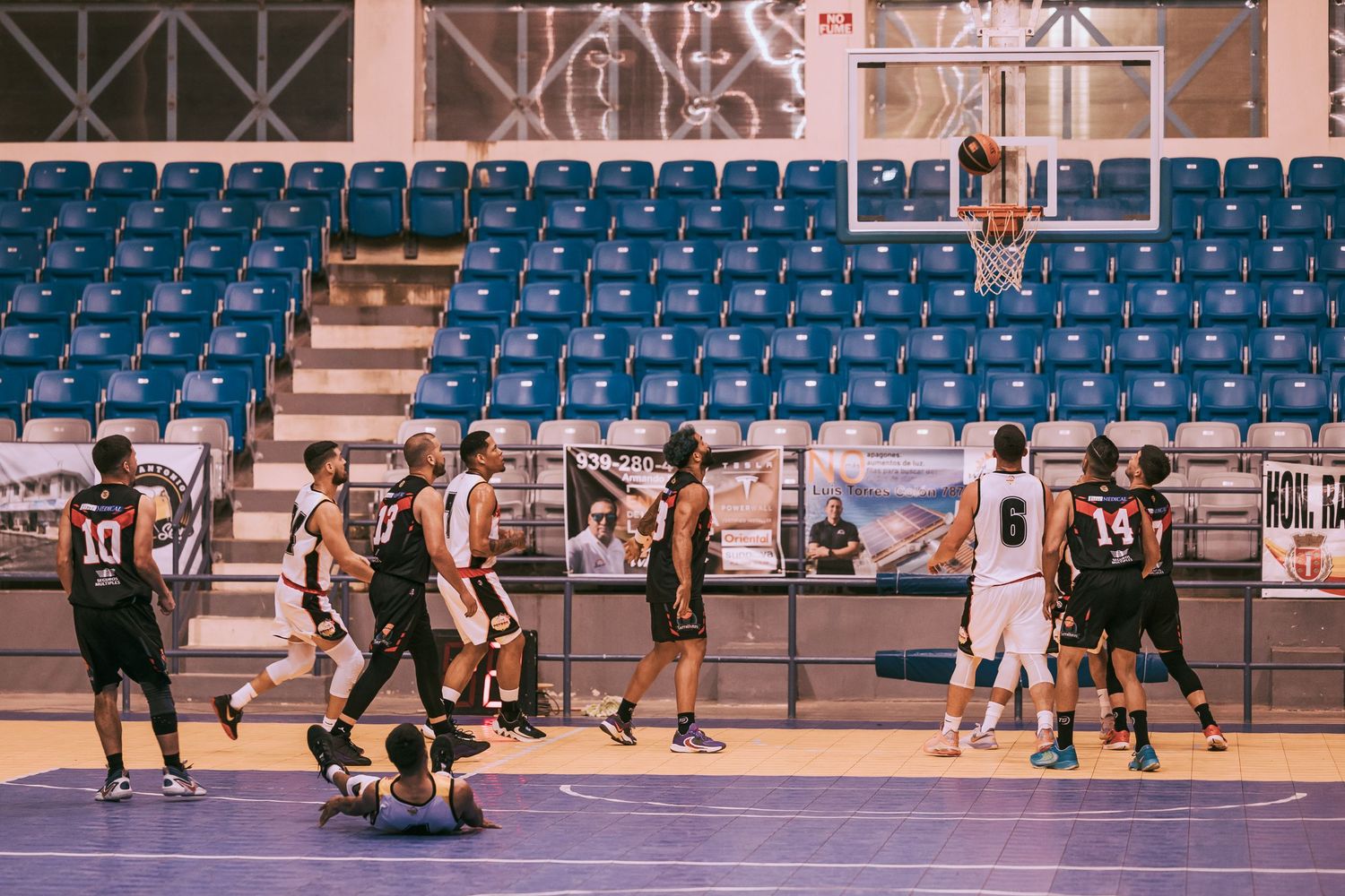 Basketball players compete on an indoor court with blue stadium seating during a game, one player near the hoop.