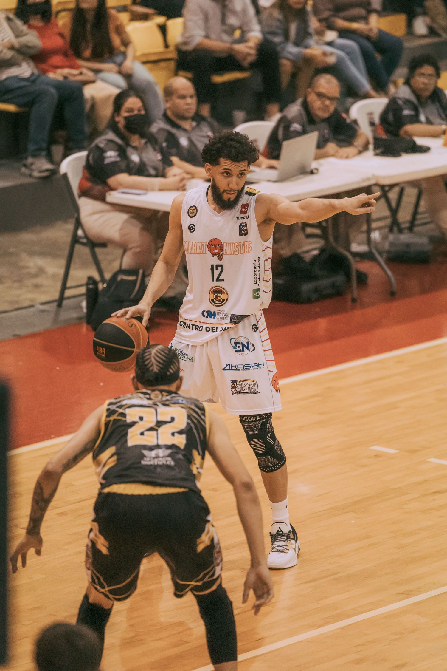 Basketball player #12 in white jersey dribbles against defender #22 during an indoor game with spectators watching.