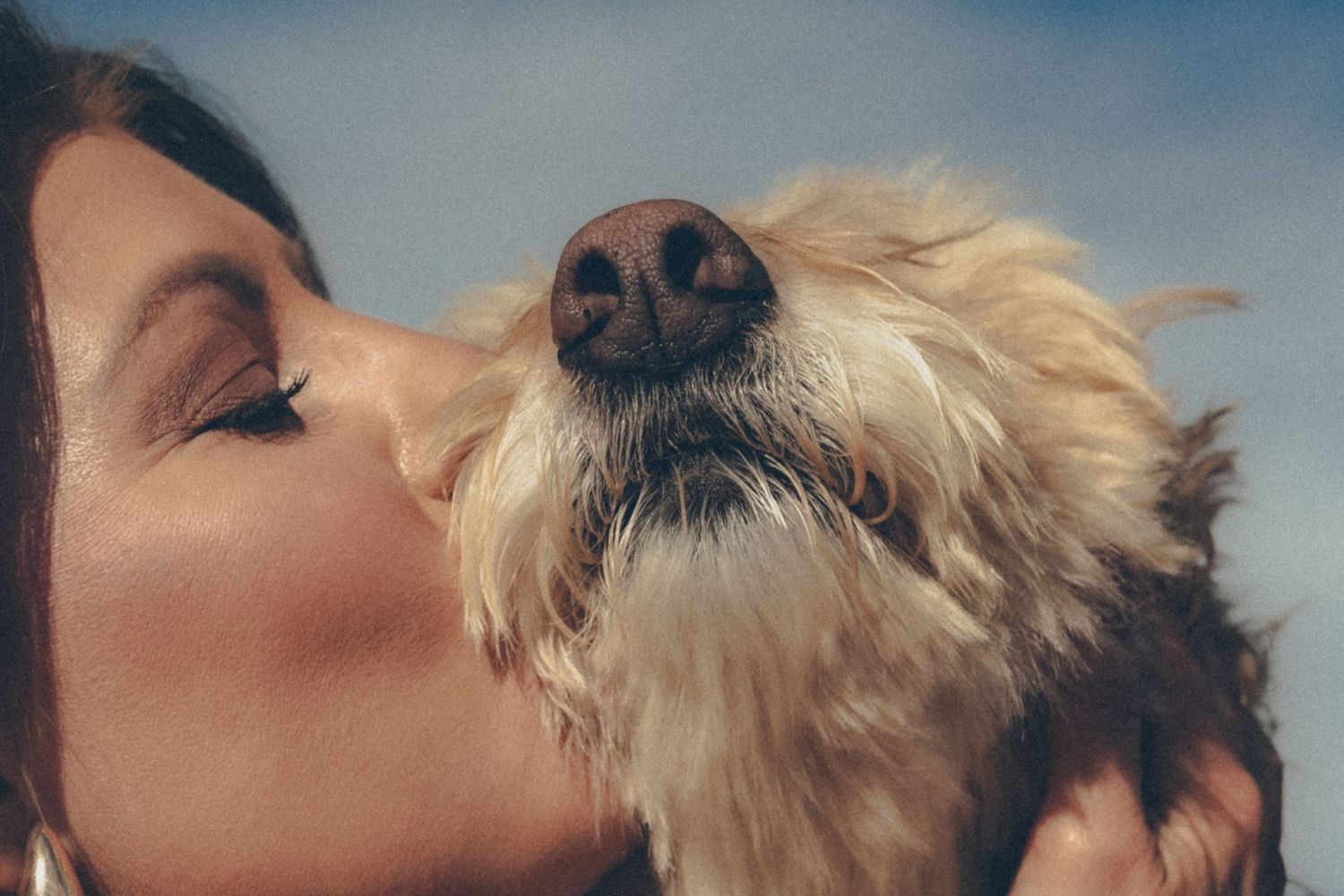 A woman with closed eyes kissing a fluffy golden dog on the cheek against a blue-sky background.