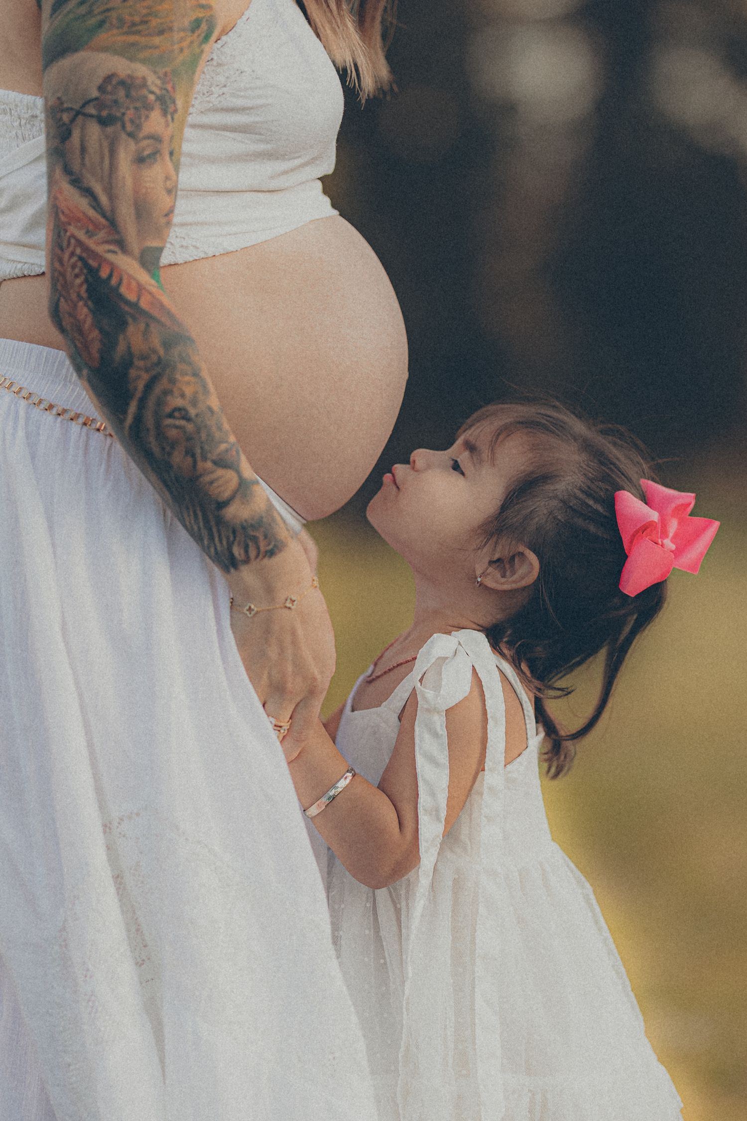 A little girl in a white dress looks up at her pregnant mother's belly outdoors.