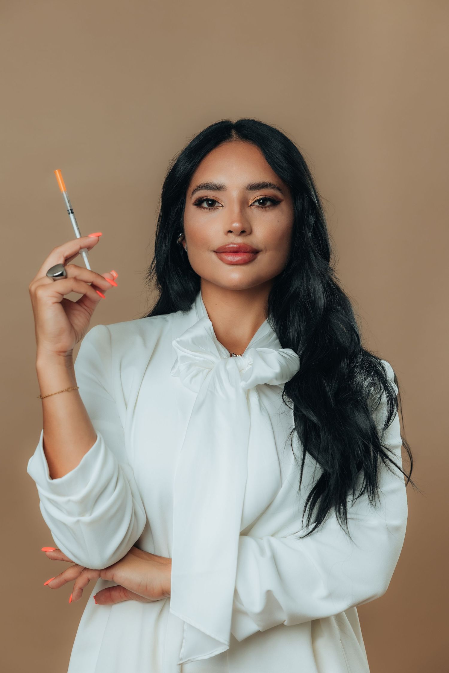Woman in white blouse holding a syringe, posing confidently against a beige background.