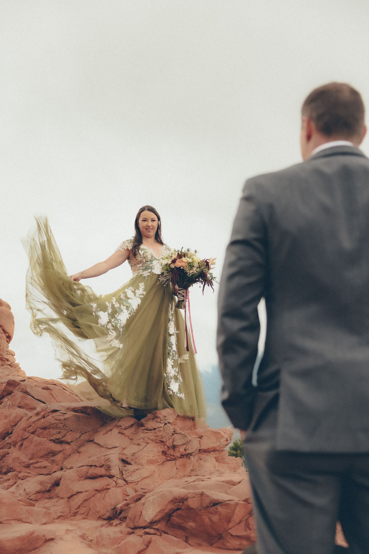 Bride in flowing olive-green gown holds bouquet on red rock formation as groom watches during outdoor elopement.