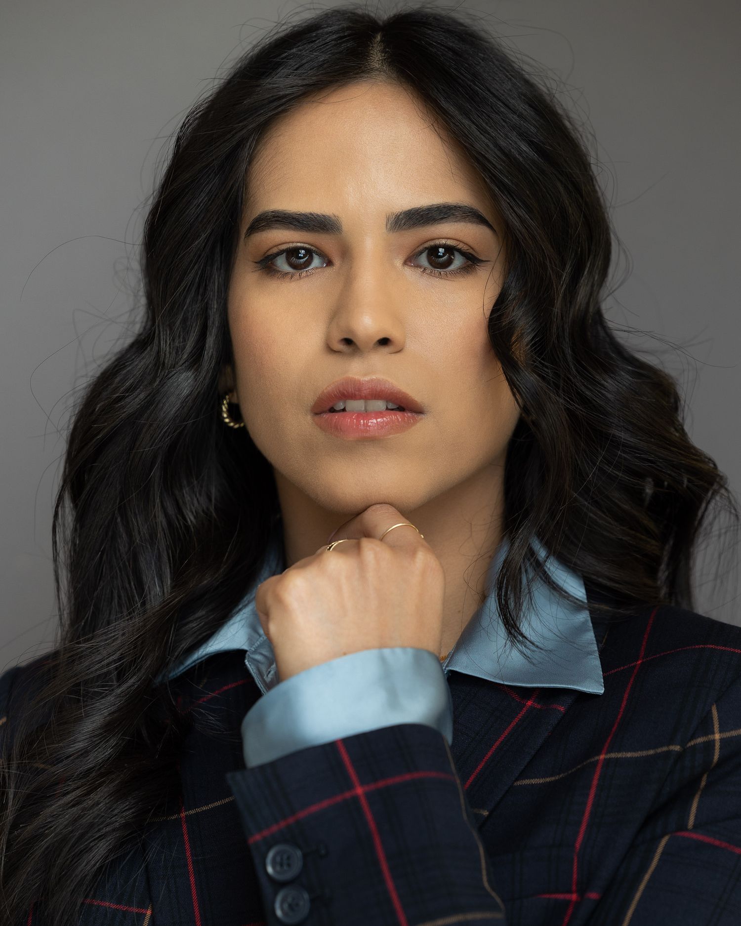 A young woman with wavy black hair poses confidently in a plaid blazer over a blue shirt against a gray background.