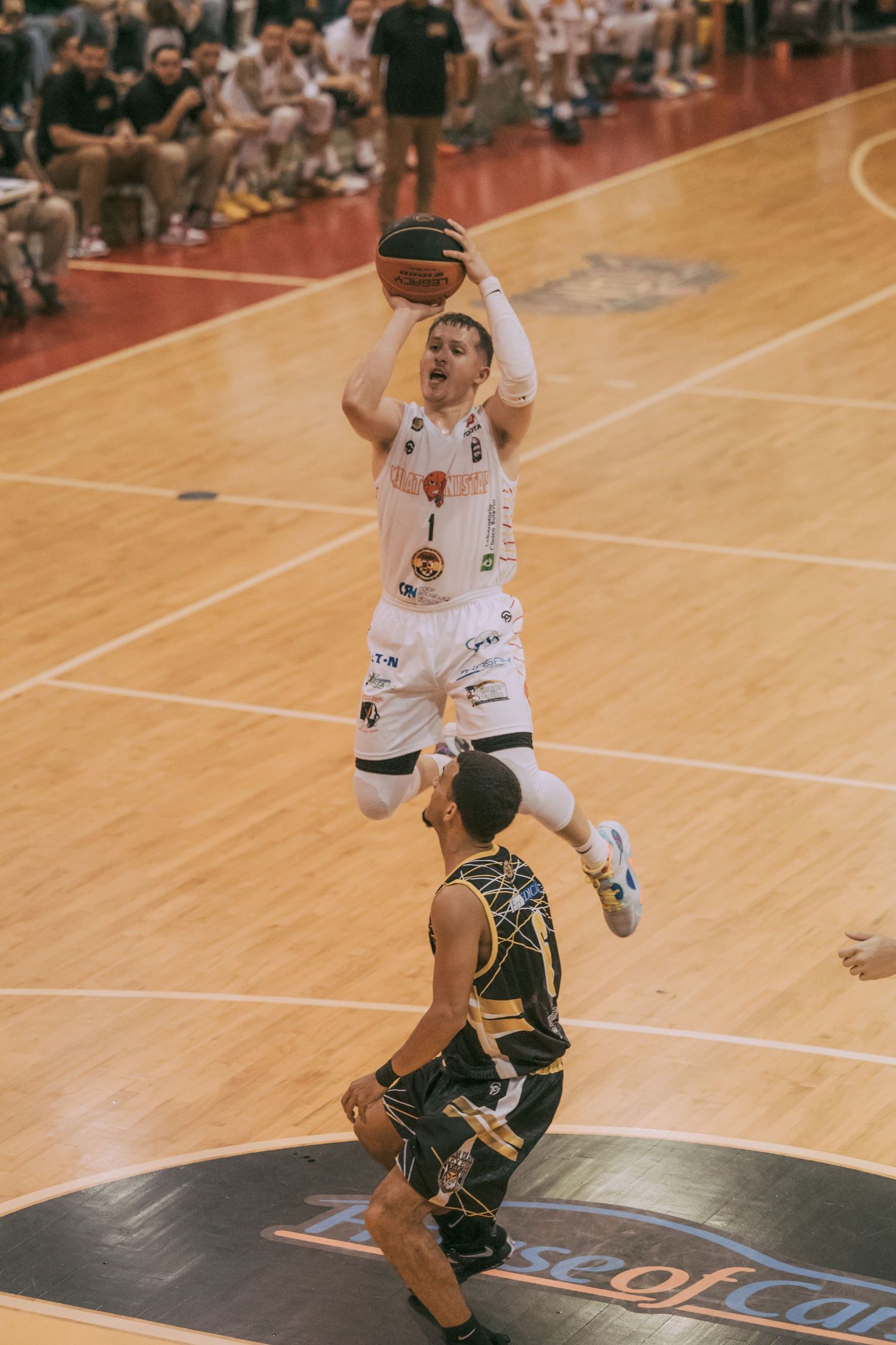 Basketball player in white uniform jumps to shoot over defender in black during an indoor game.