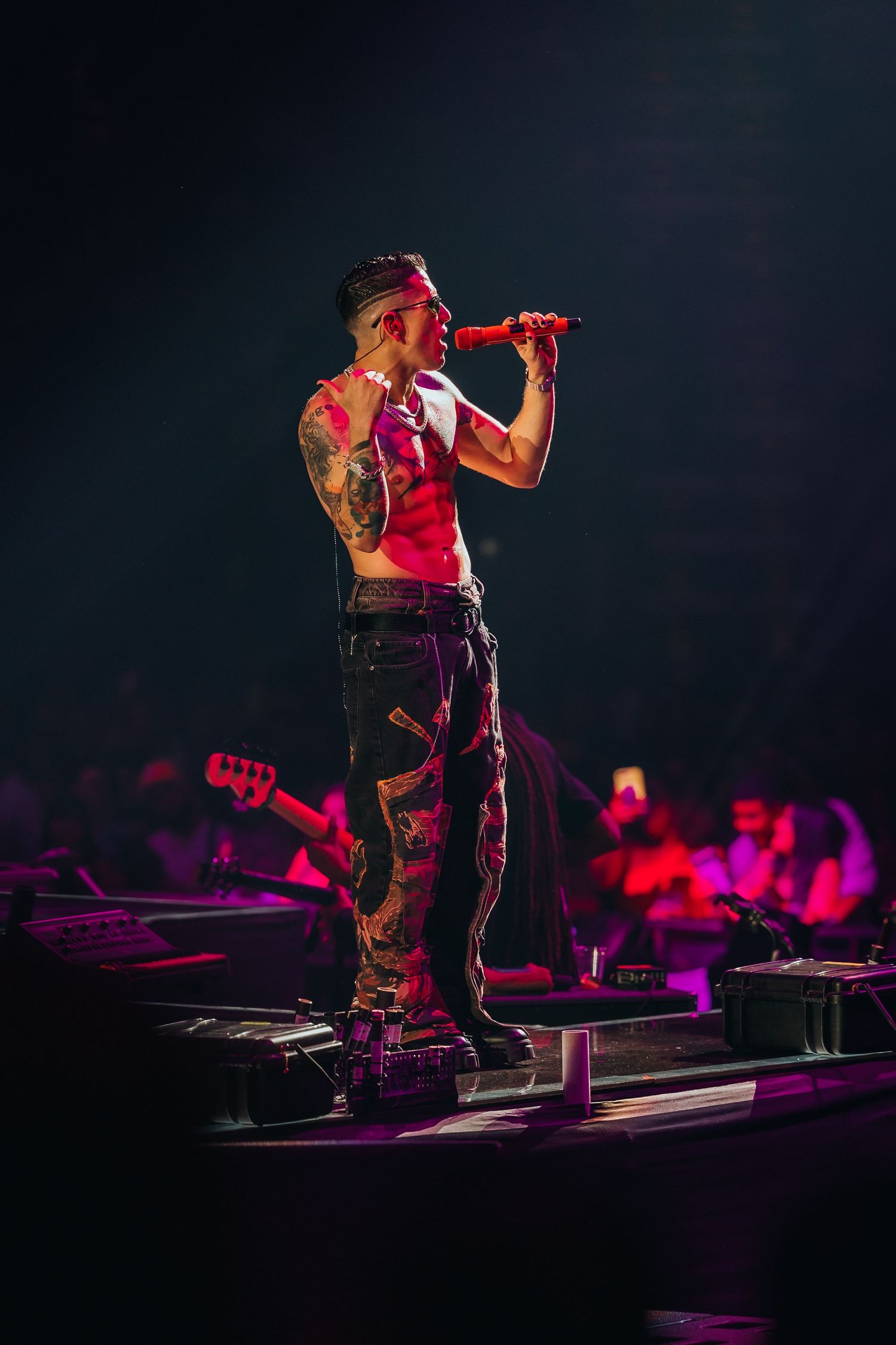 Shirtless tattooed male performer singing into red microphone on dark concert stage wearing cargo pants.