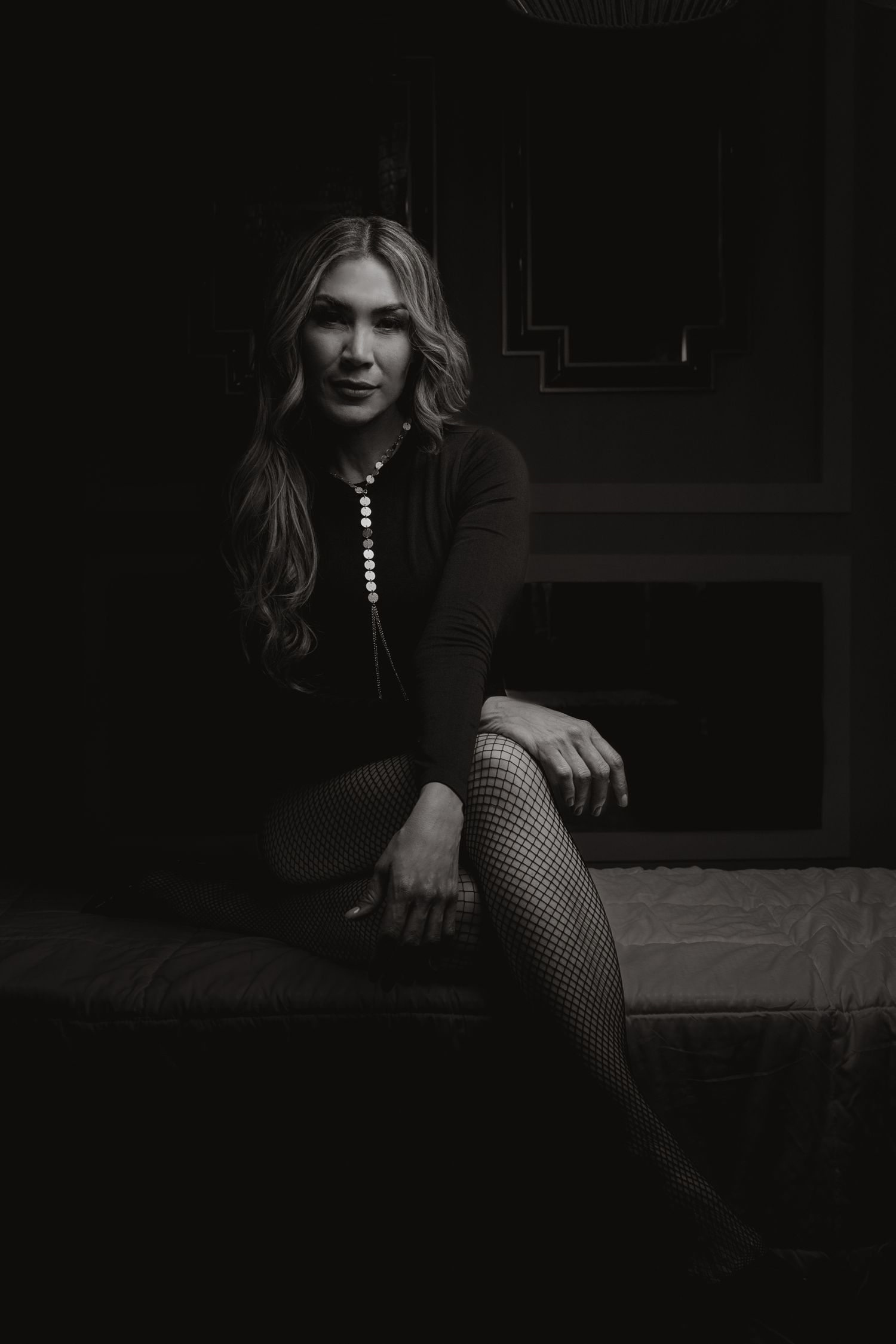 Black and white portrait of a blonde woman in fishnet tights and dark top seated in a dimly lit room.