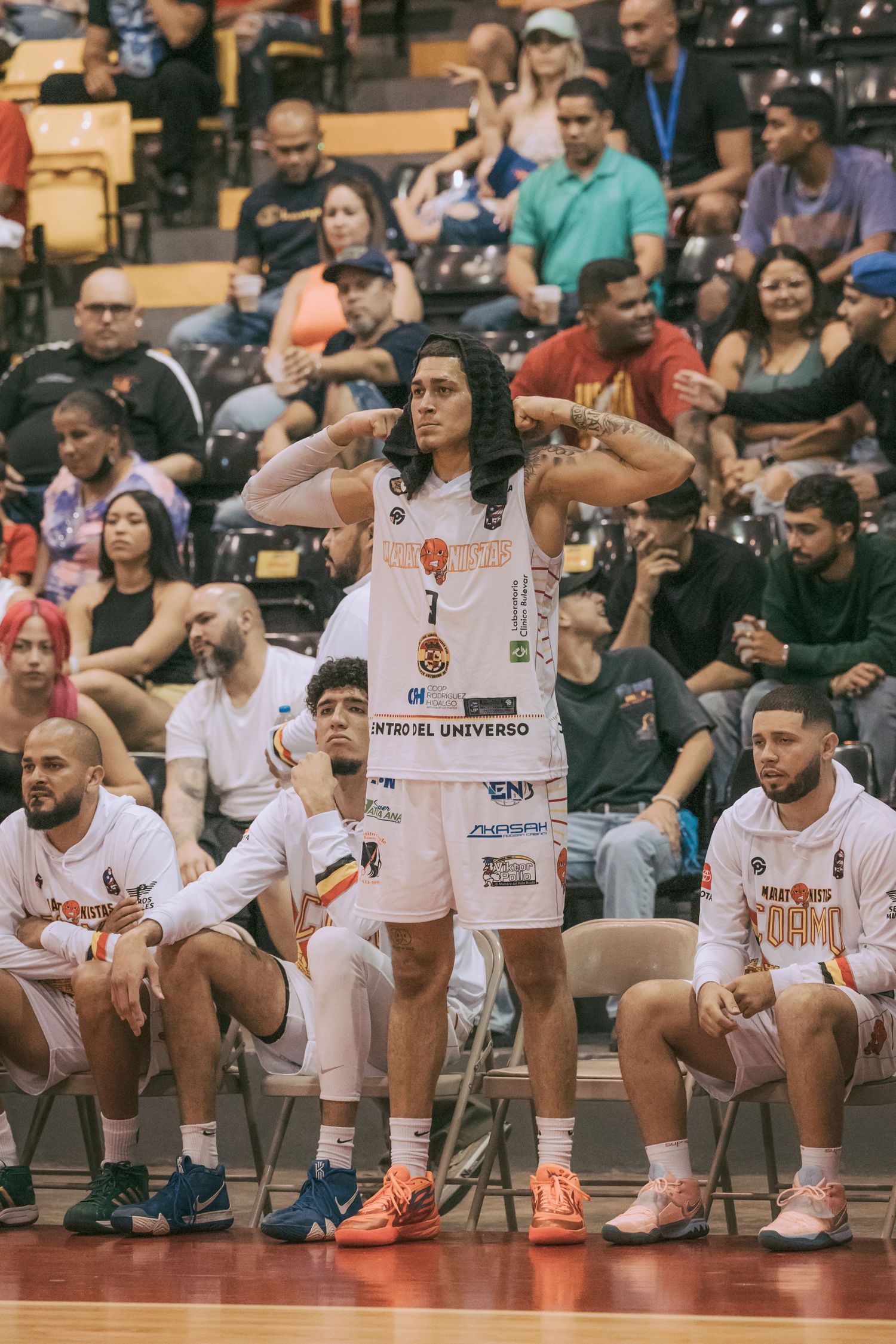 Basketball player in white jersey stands confidently on sideline during a crowded indoor game.