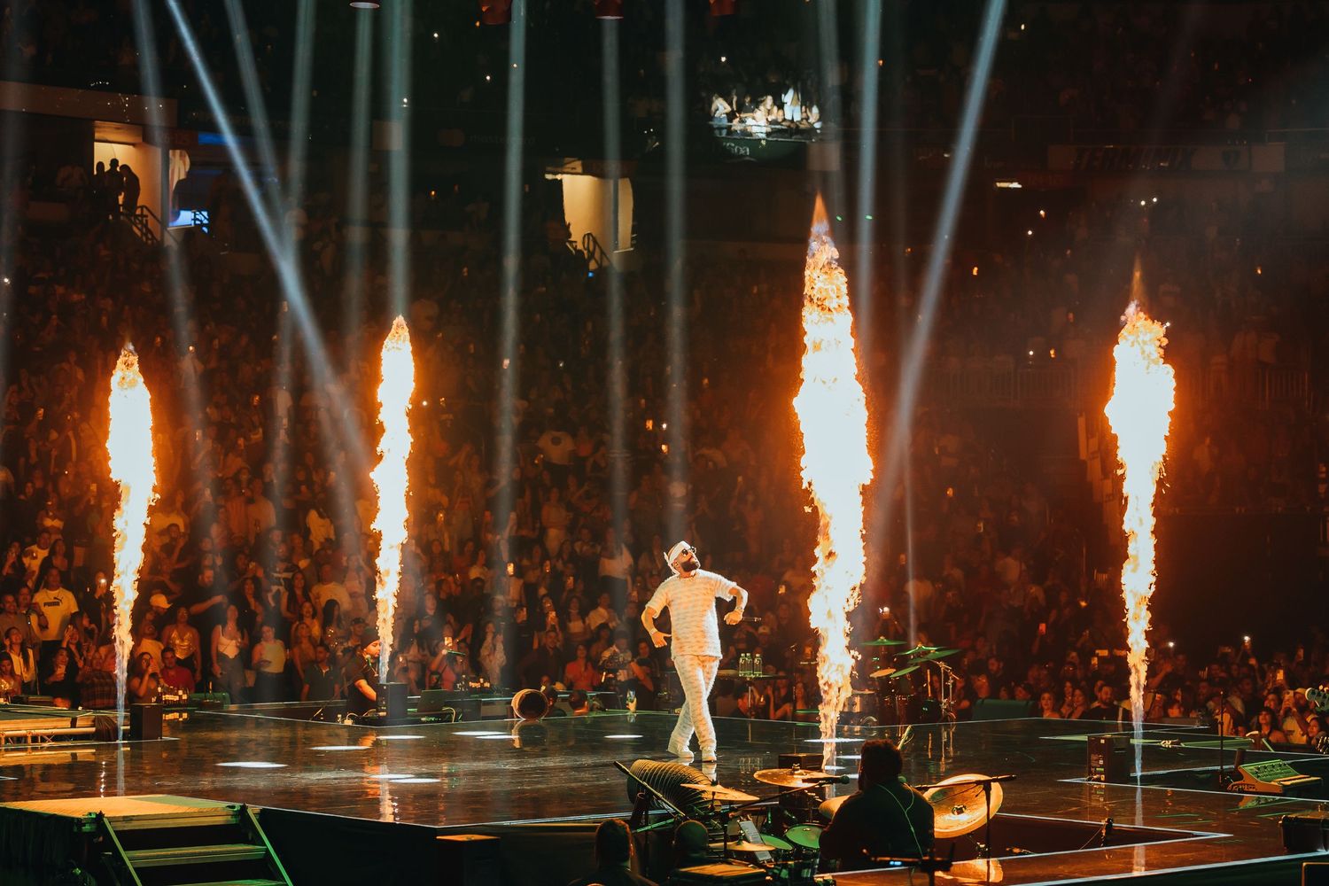 Performer on stage surrounded by massive fire columns and dramatic light beams at a packed concert arena.