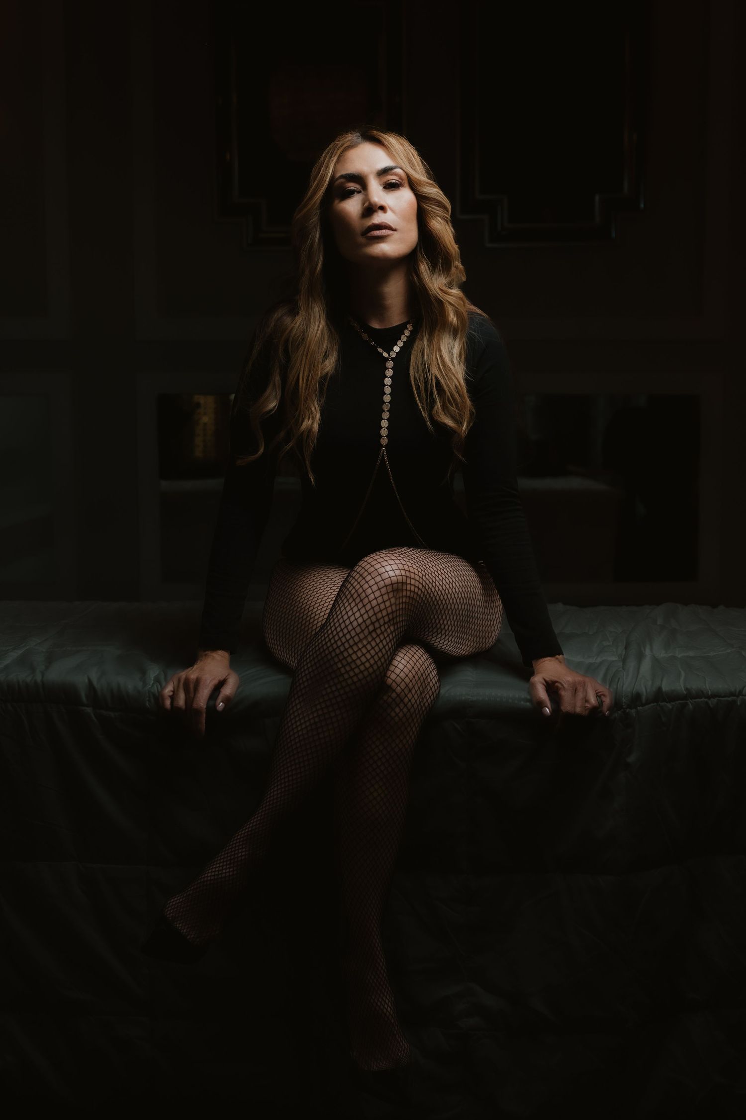 Woman in black outfit and fishnet stockings sitting on a dark bed with moody dramatic lighting.