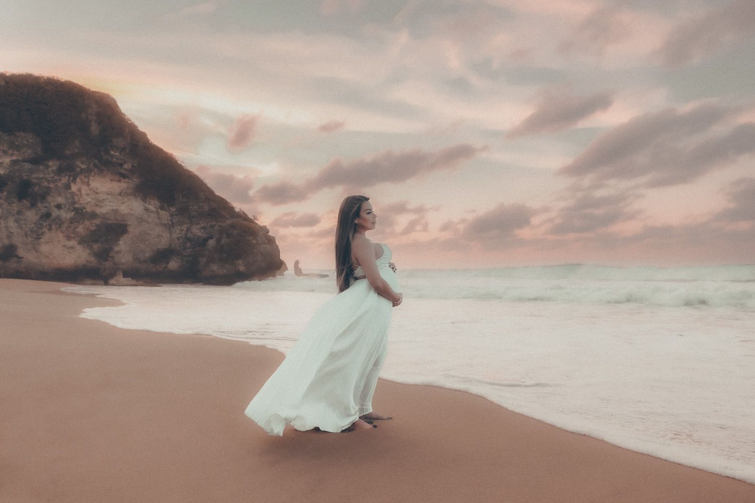 Expecting mother in a flowing white maternity dress standing on a beach at sunset