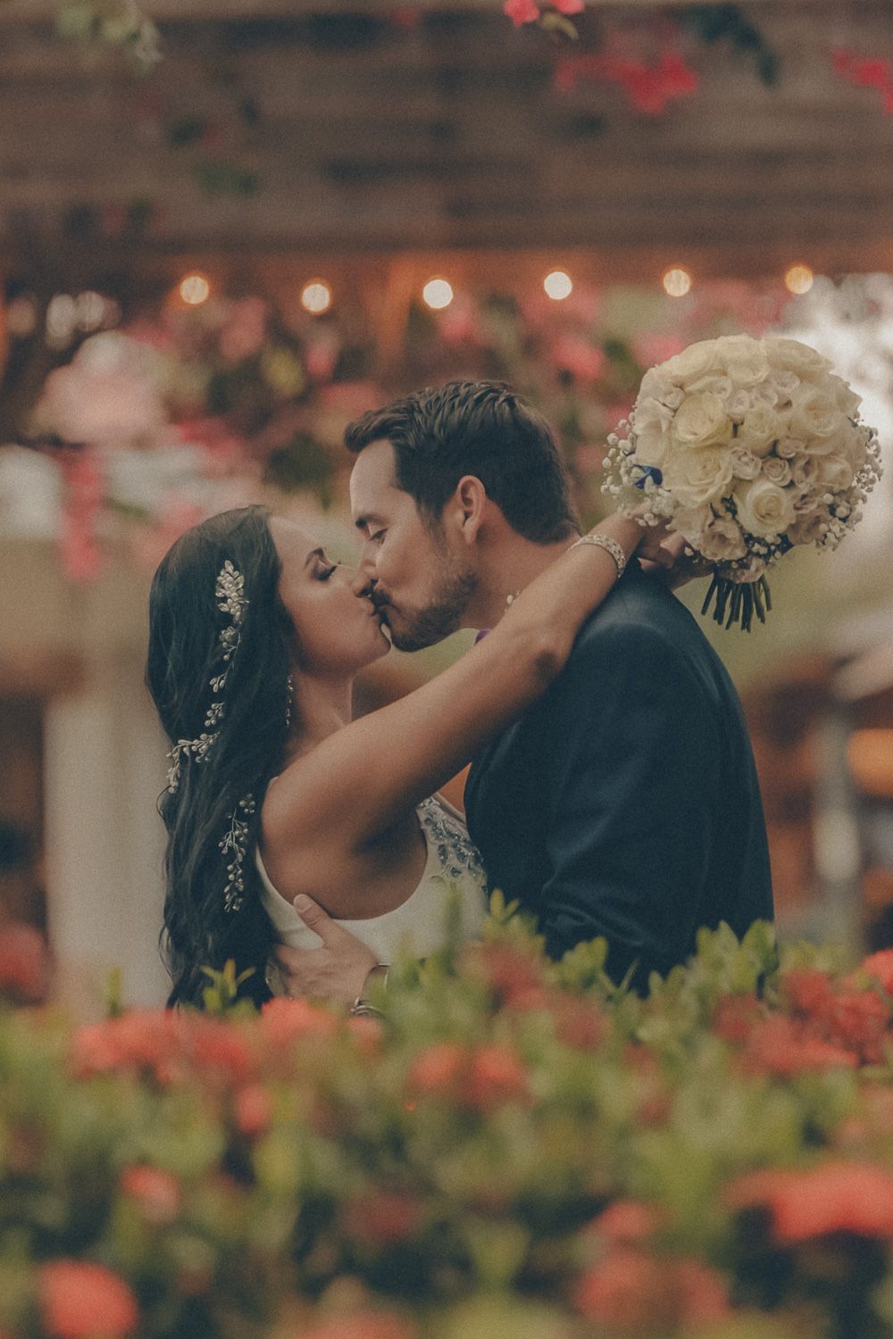 A bride and groom share a romantic kiss surrounded by colorful flowers and string lights at an outdoor wedding venue.