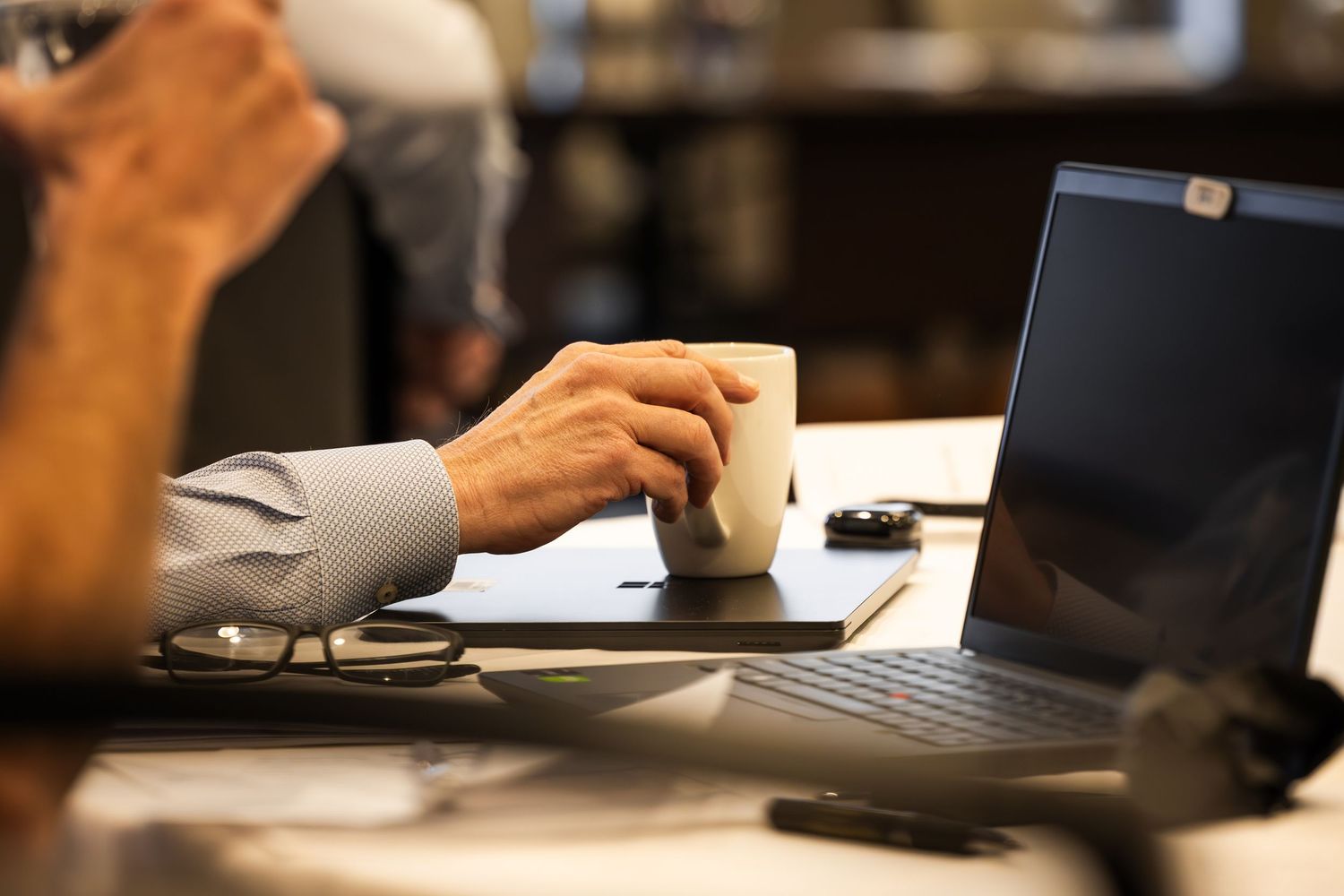 Businessman in blue shirt holding coffee cup at desk with open laptop and glasses in office setting.