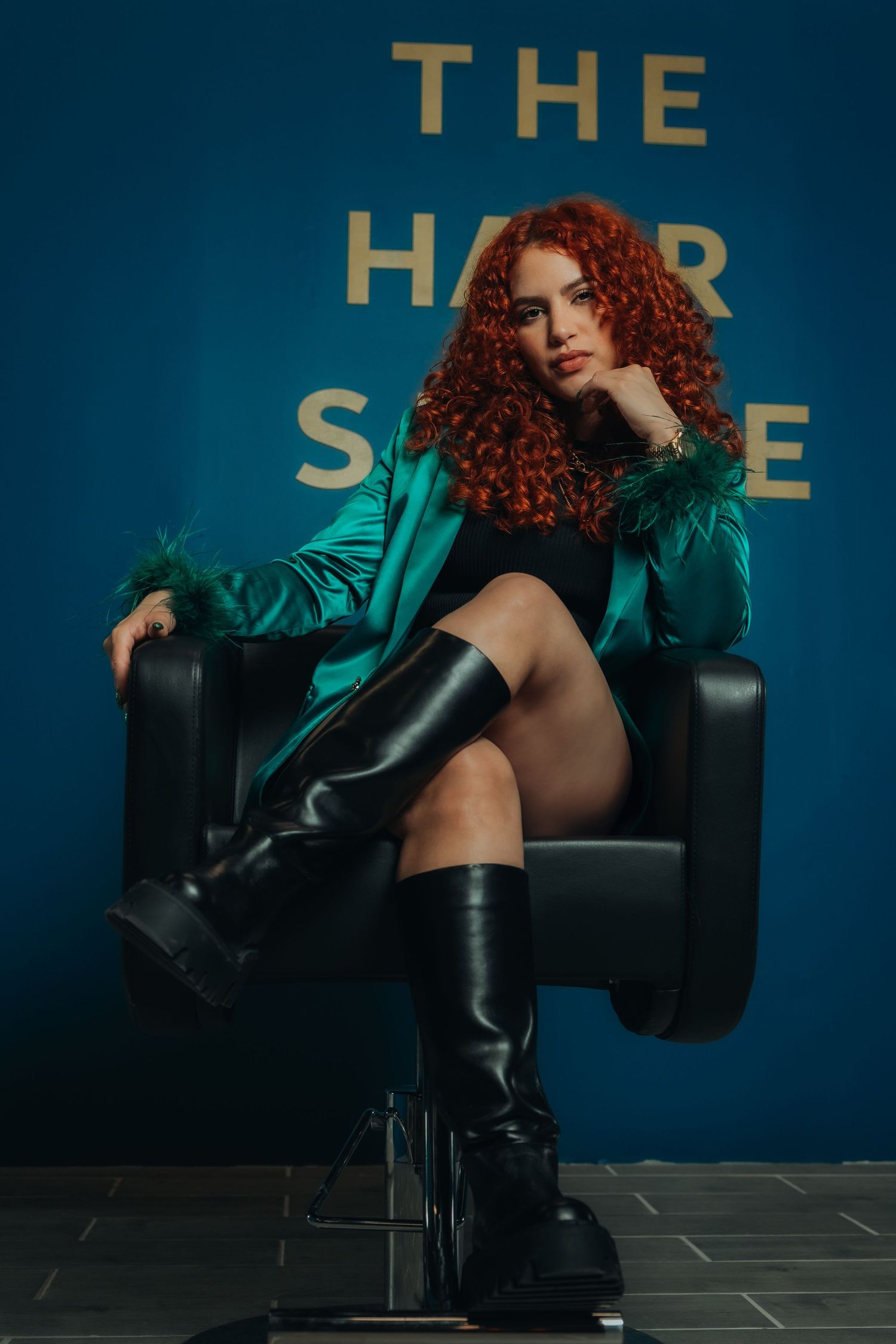 A woman with curly red hair sits in a salon chair wearing a green jacket and black knee-high boots at The Hair Salon.