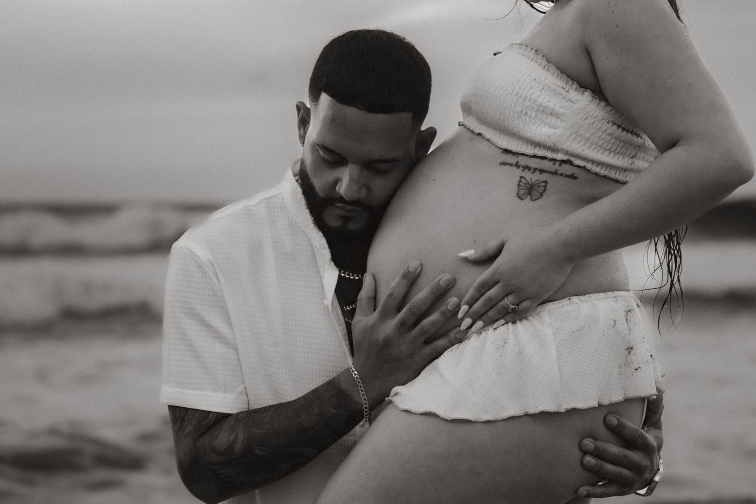 Black and white maternity portrait of an expecting father embracing his partner's baby bump on the beach