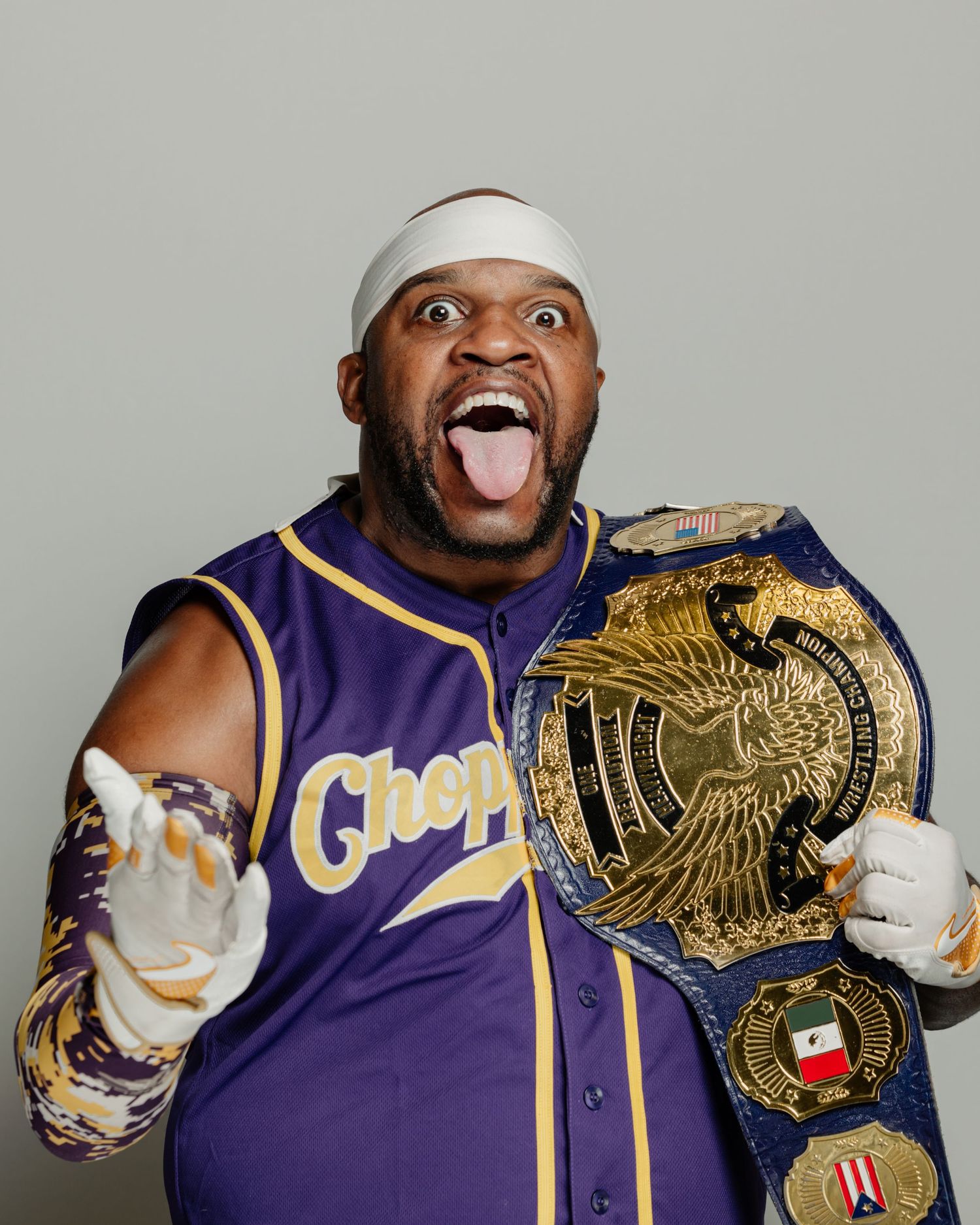 Excited man in purple Chop jersey holds championship wrestling belt, wearing white gloves and headband, tongue out.