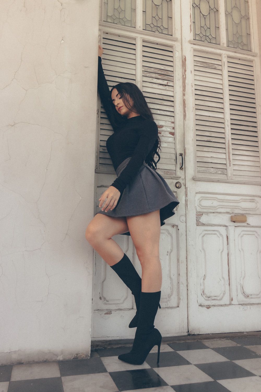 Full-body fashion portrait of brunette model in black outfit and knee-high boots beside vintage doors