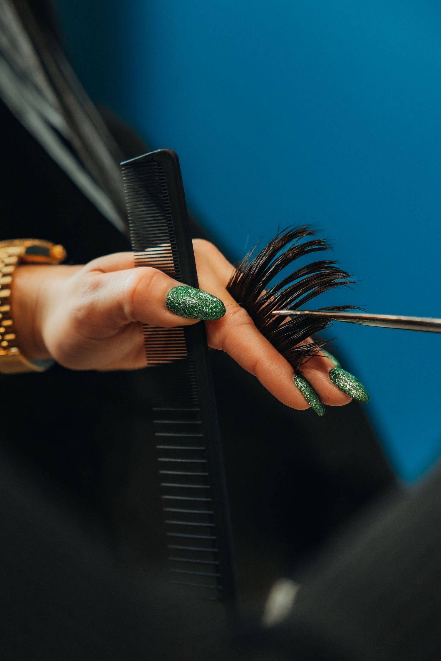 Hairstylist with green glitter nails cutting dark hair with scissors and comb in a salon.