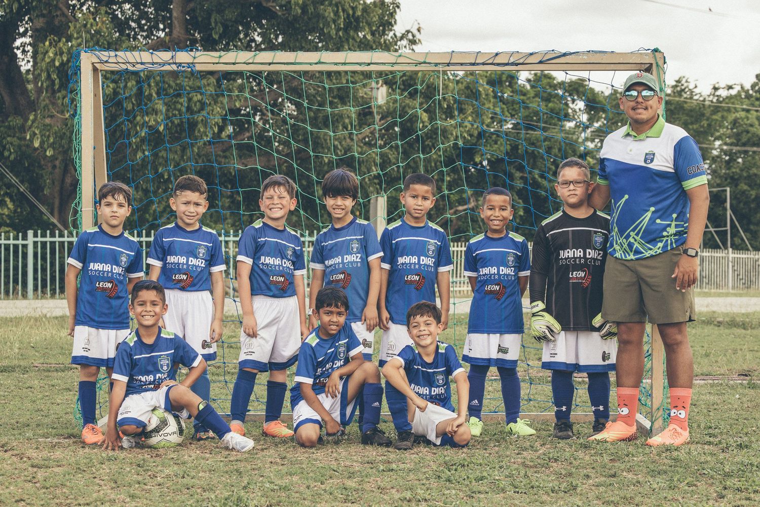 Young boys soccer team in blue uniforms posing with coach in front of goal on green field.