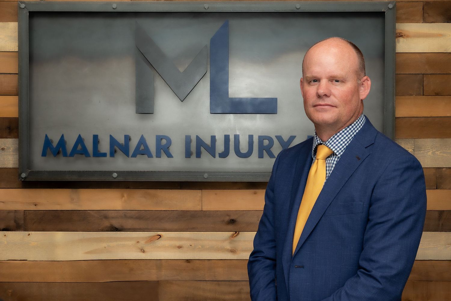 Professional headshot of a man in a suit standing before the Malnar Injury Law firm sign with wooden wall backdrop.