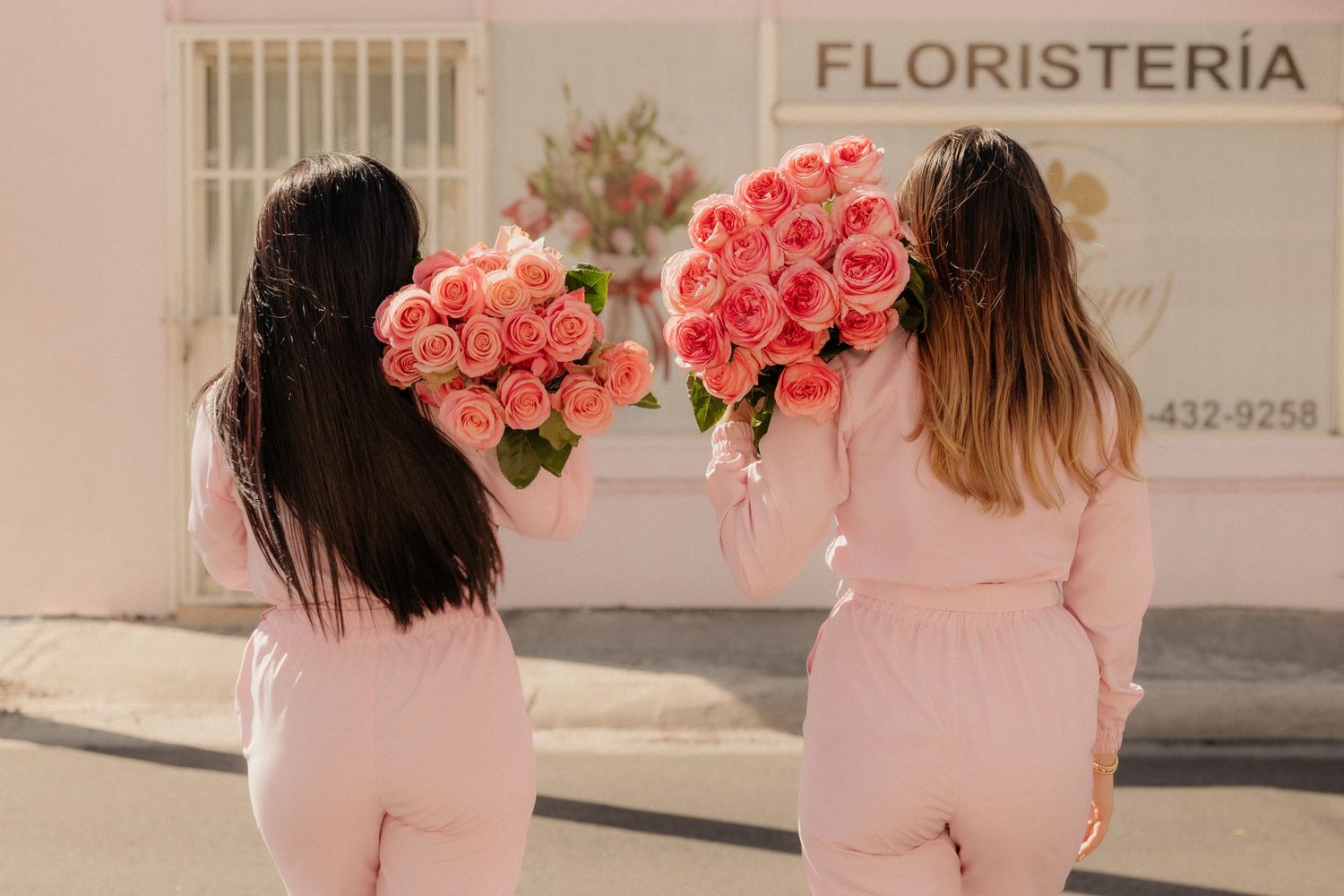 Two women in pink outfits hold large bouquets of pink roses outside a flower shop.