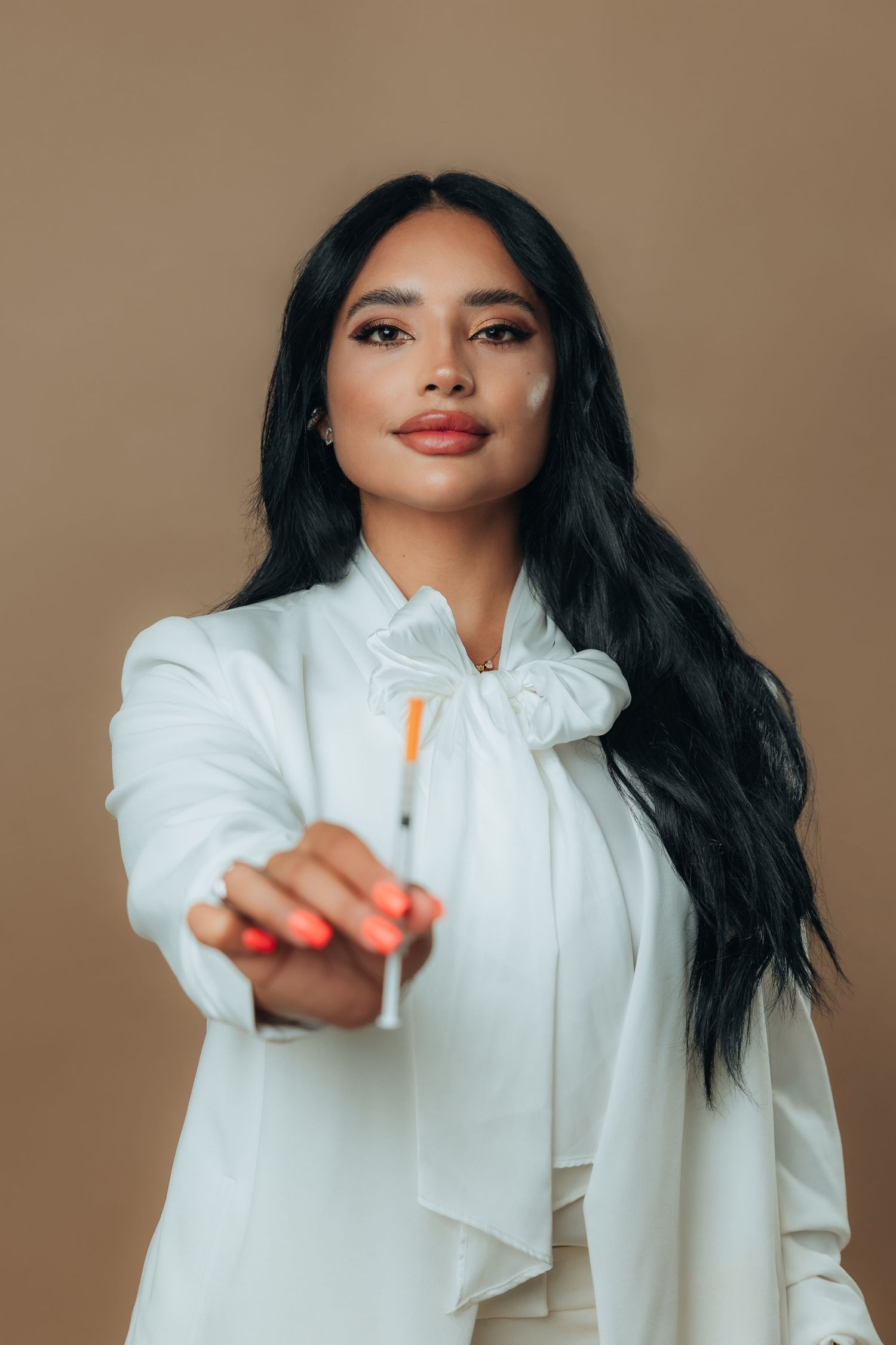 Woman in white blouse holding a syringe toward camera against beige background.