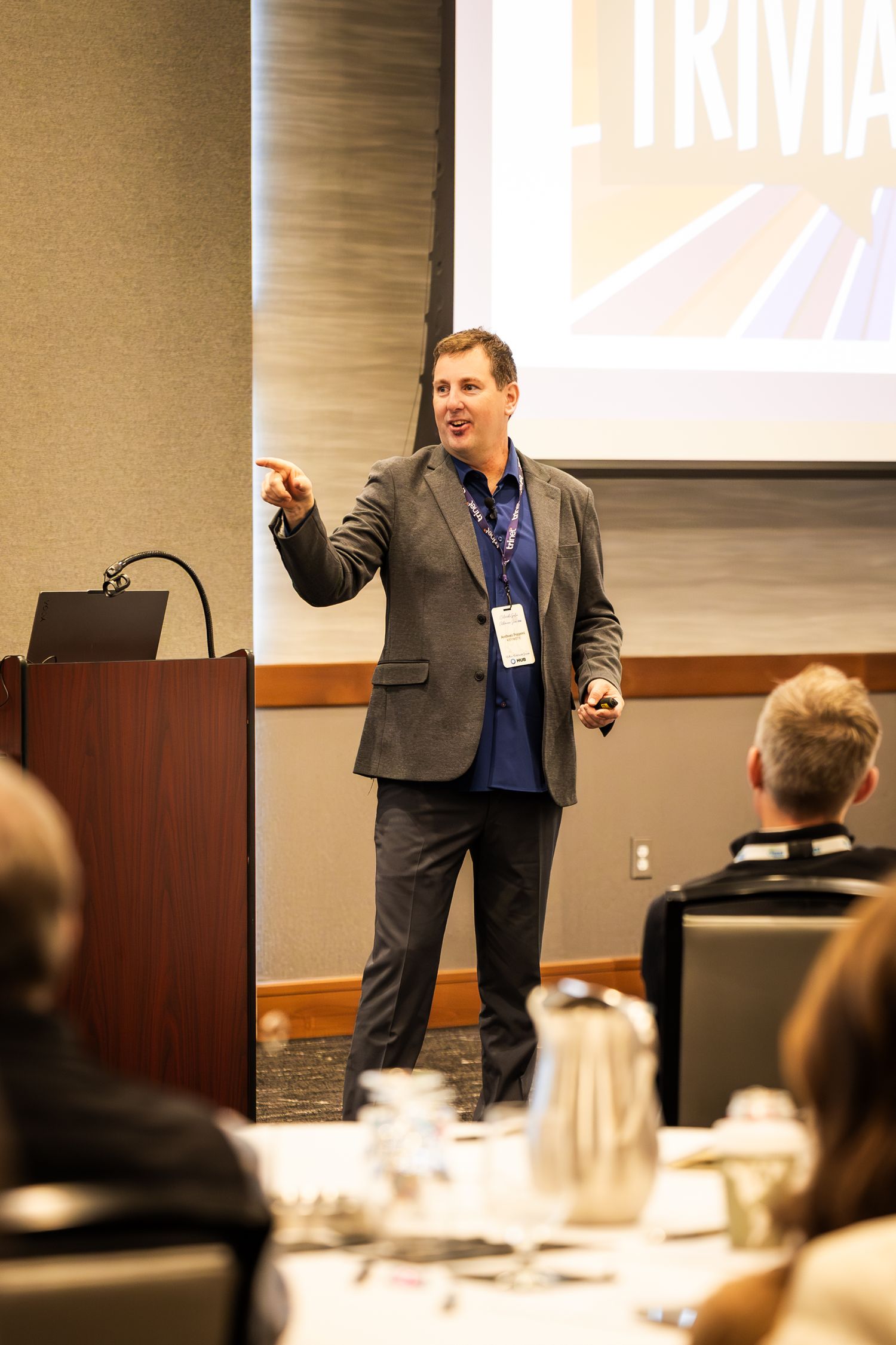 A male speaker presents at a conference, pointing at a trivia slide on a projector screen in a hotel ballroom.