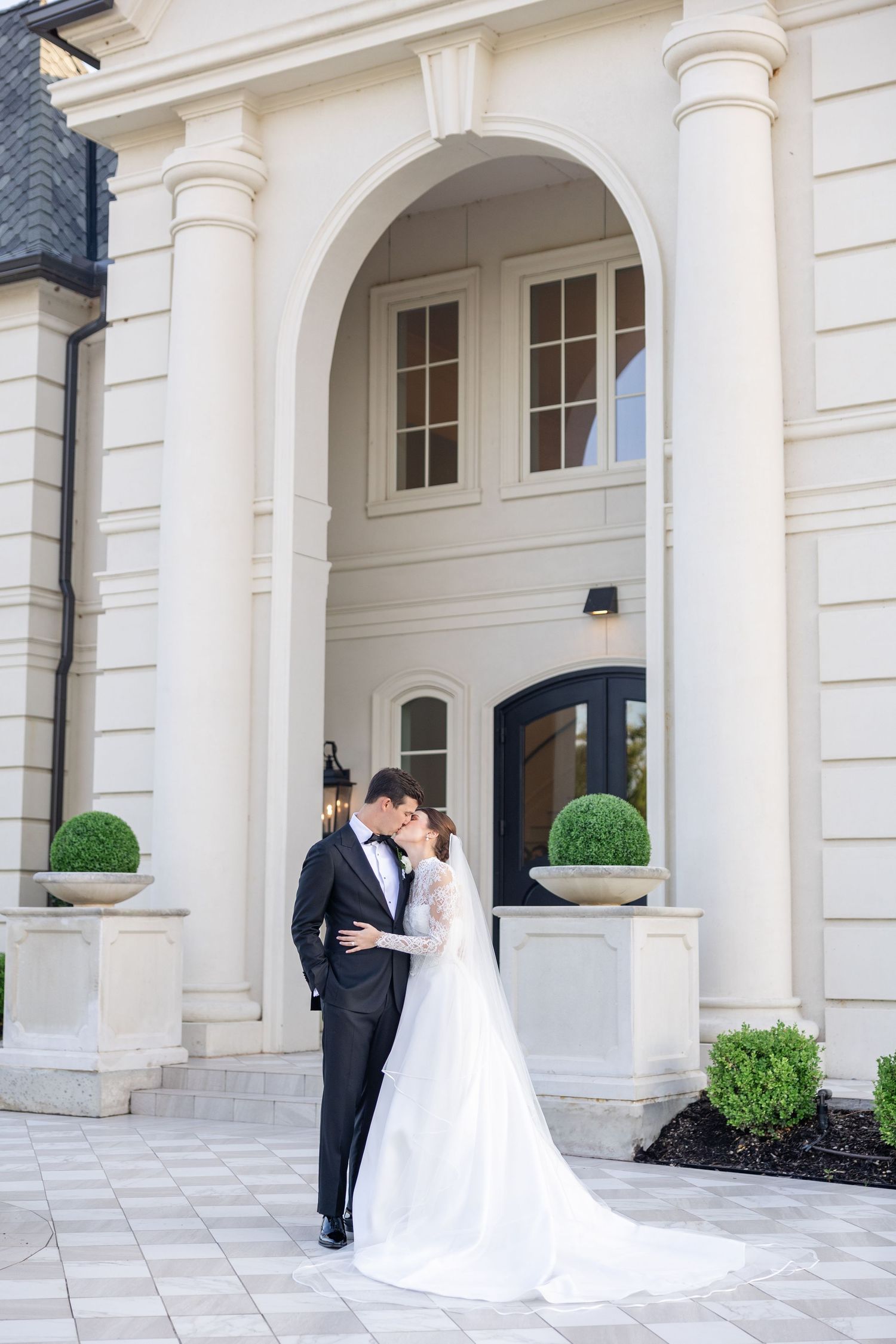 Bride and groom embracing under an elegant white arched portico of a grand estate wedding venue.