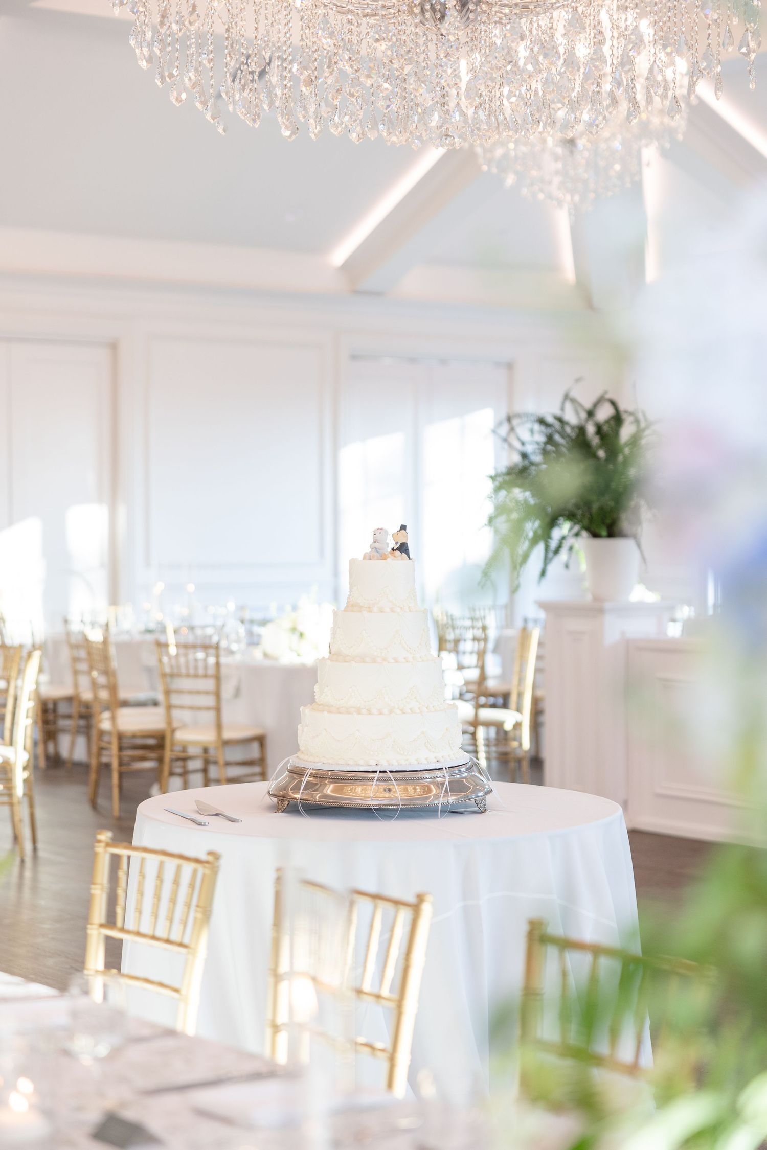Four-tier white wedding cake with couple topper displayed on round table beneath crystal chandelier in bright ballroom.