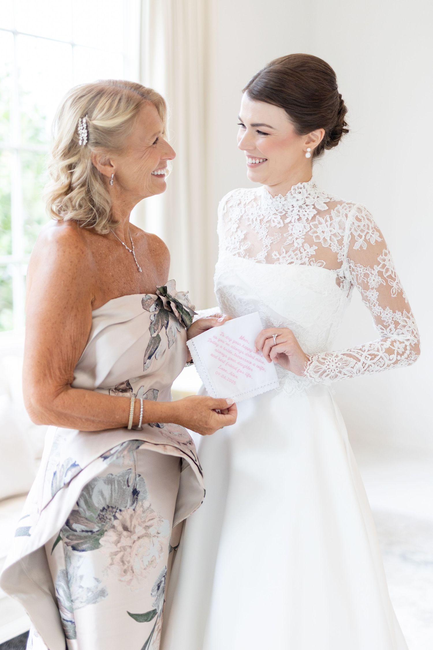 Smiling bride in high-neck lace gown sharing handkerchief note with mother in floral dress.