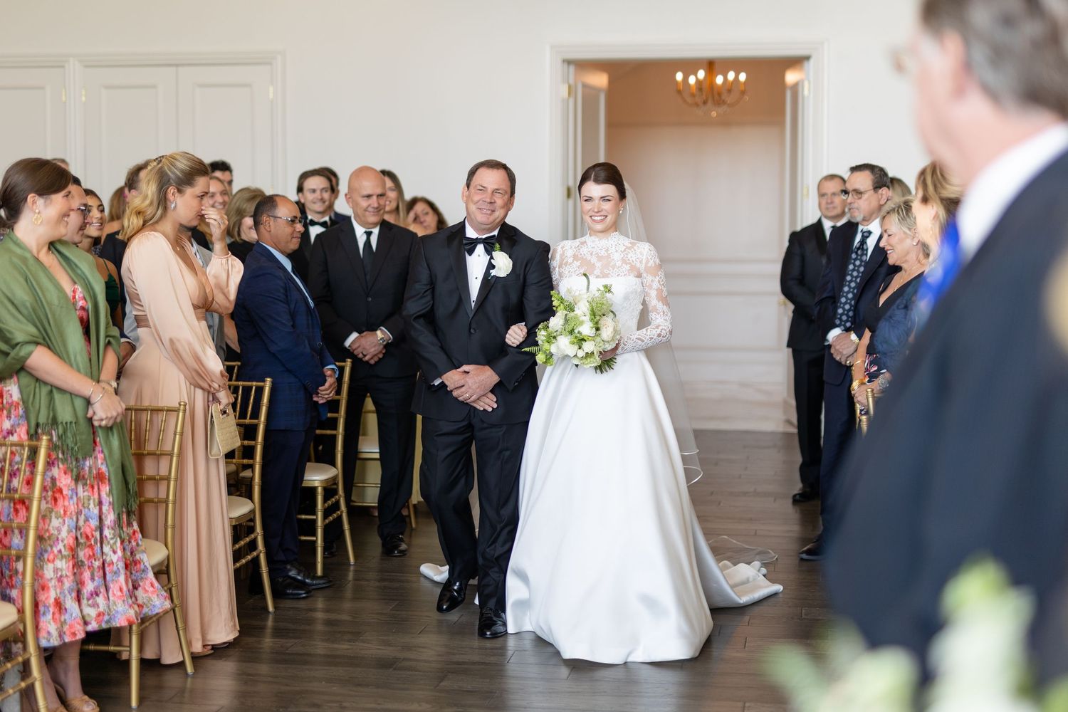 Bride in lace wedding gown walks down the aisle with her father at an elegant white venue.