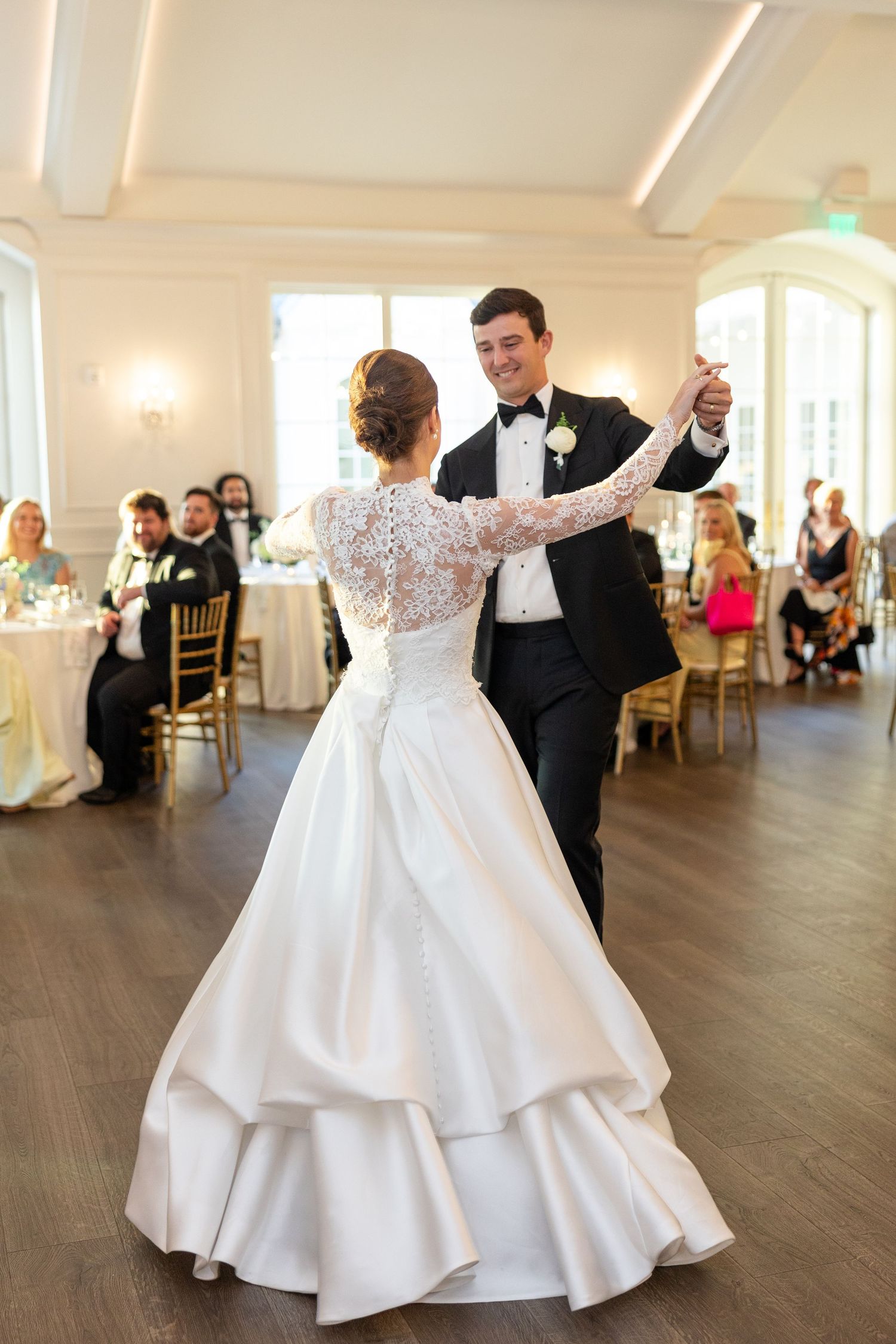 Bride in lace long-sleeve gown and groom in tuxedo share romantic first dance in bright white ballroom.